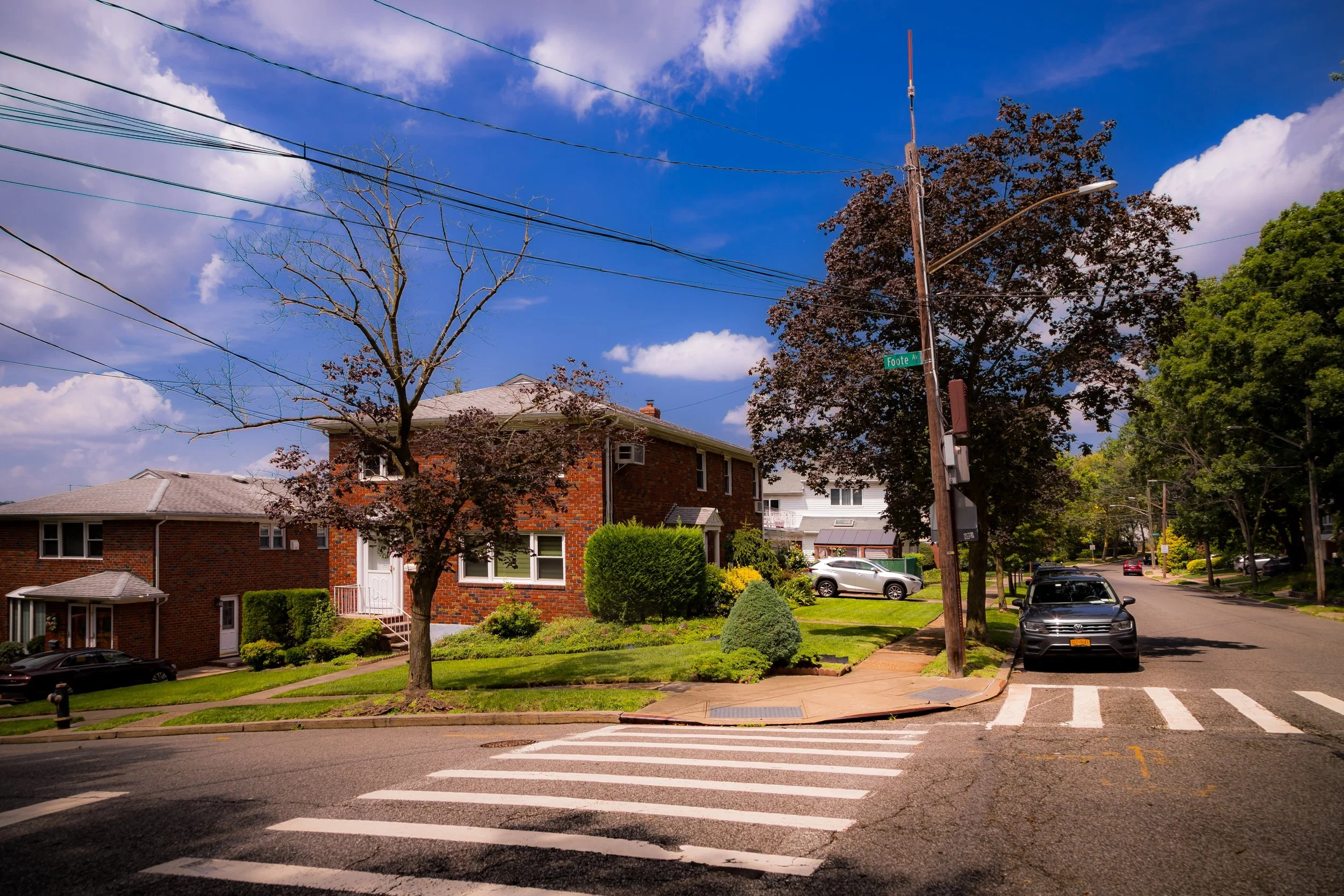 A suburban neighborhood street corner on a sunny day with a brick house, trees, parked cars, a crosswalk, and utility poles.