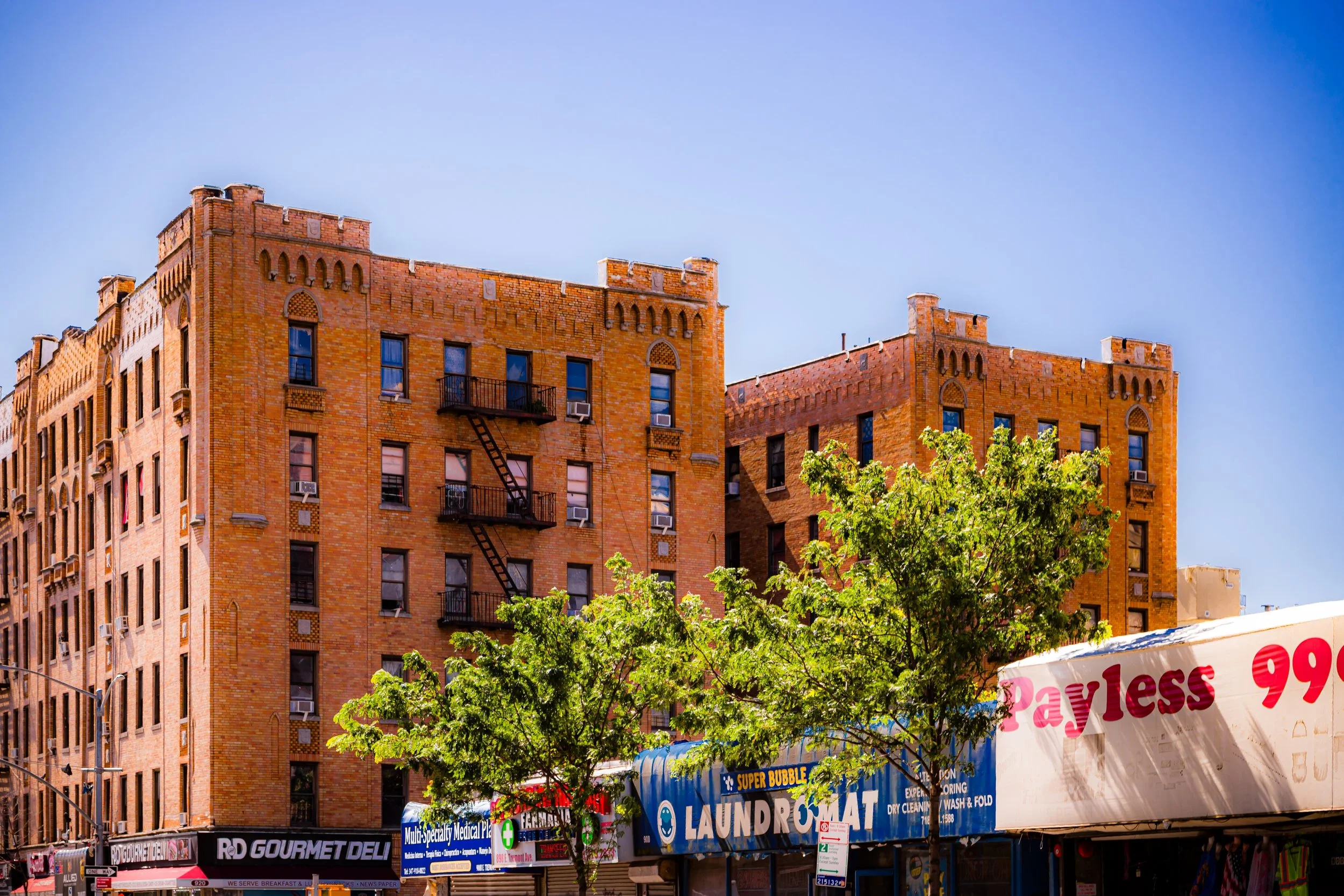 A city street scene featuring a tall brick apartment building with fire escape ladders and a bright blue sky. The street level has storefronts, including a deli, laundromat, and a store with a payless sign. There are green trees in front of the buildings.