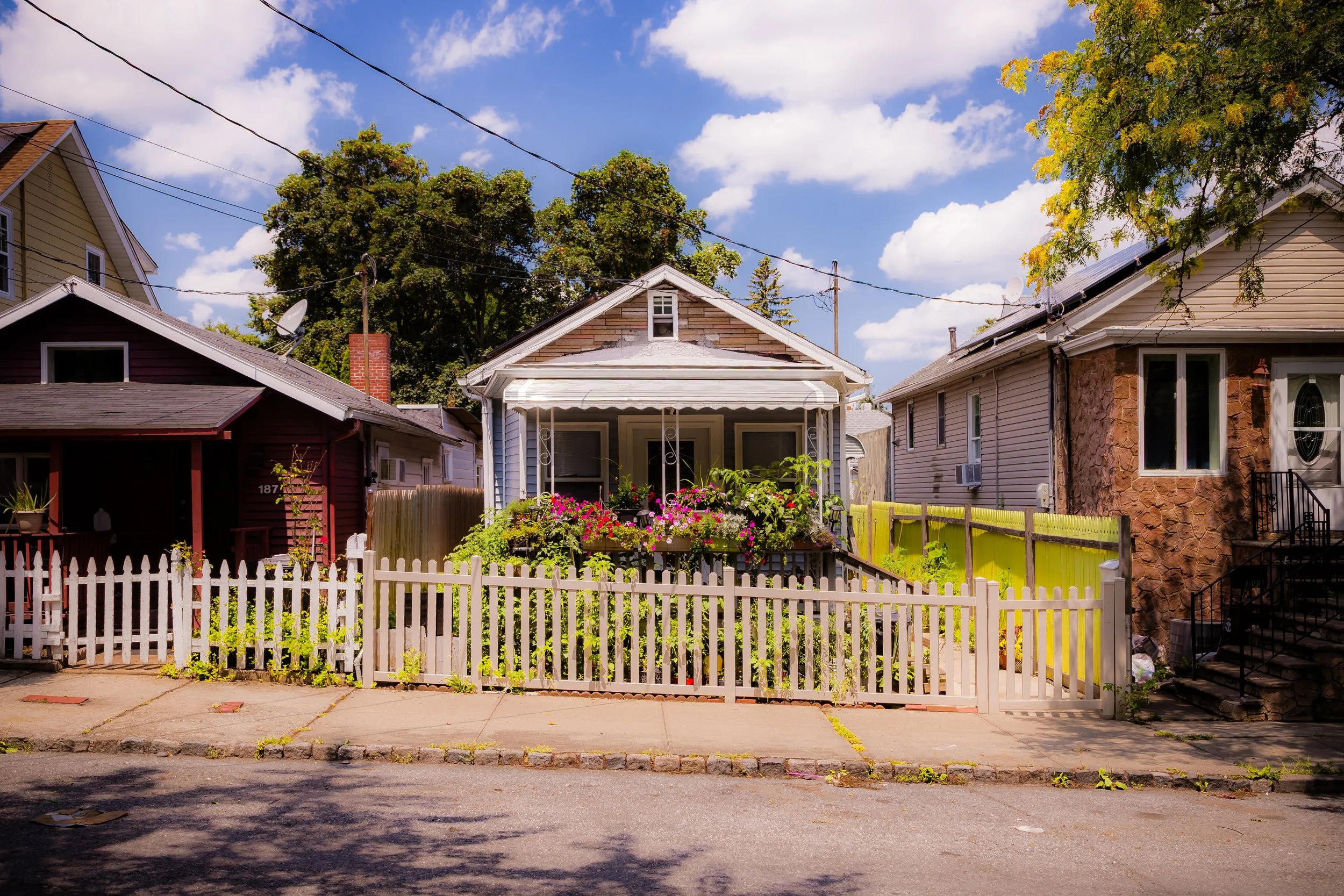 Front view of a small house with a white picket fence, surrounded by colorful flowers and greenery under a partly cloudy sky.