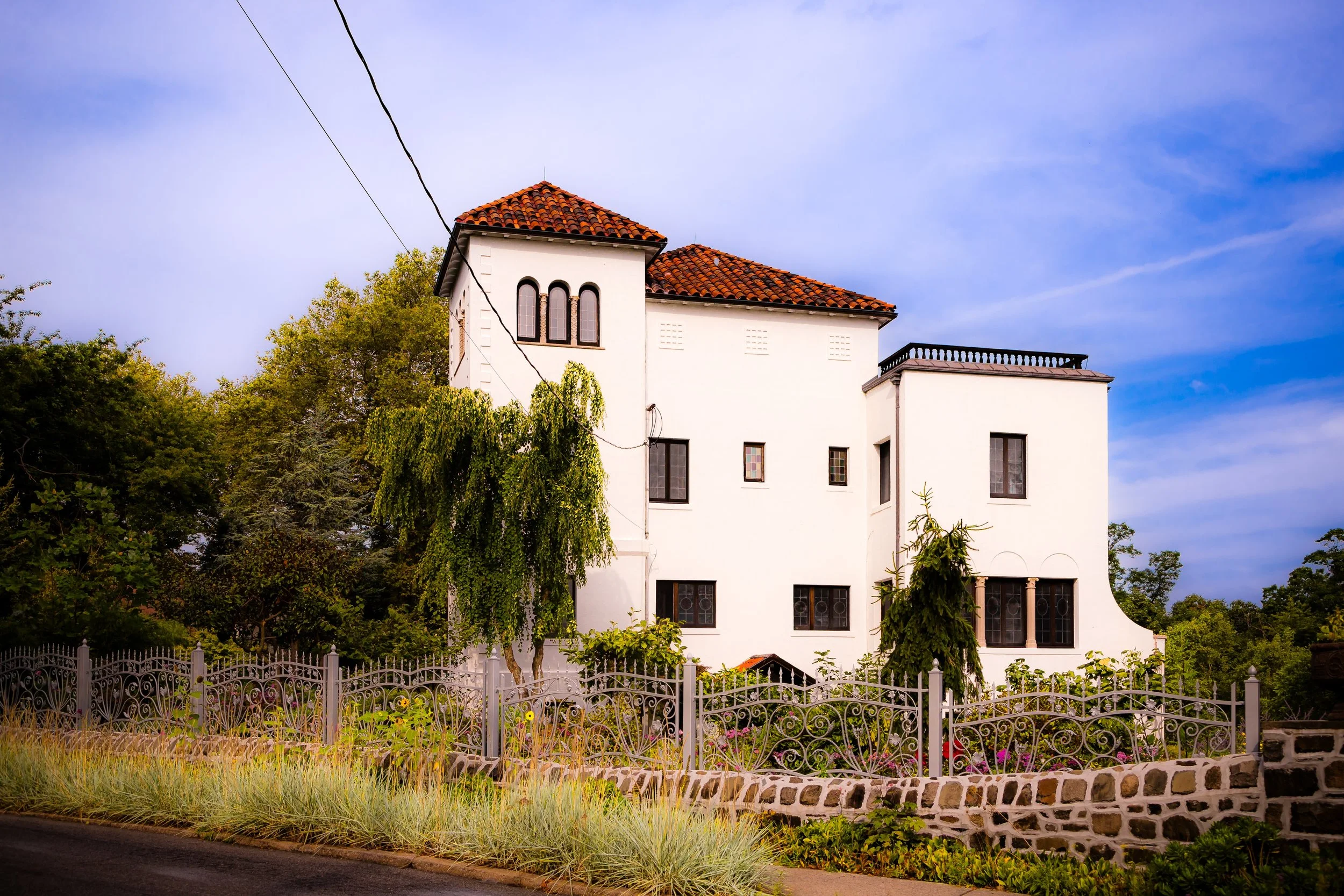 A white multi-story house with a red tile roof, surrounded by trees and decorative iron fencing, under a blue sky with wispy clouds.