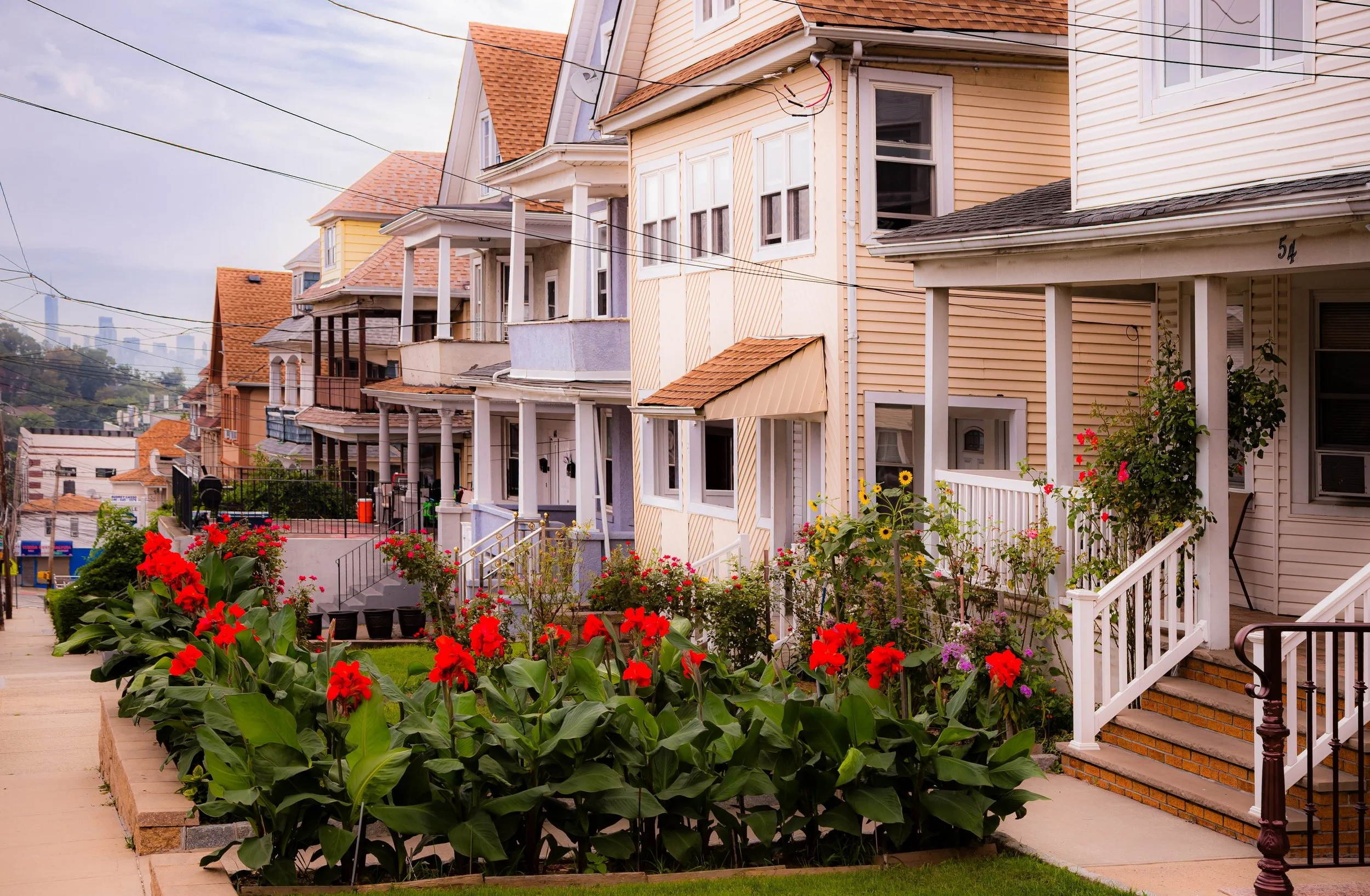 Colorful row of Victorian houses and flowers on a sidewalk in a hilly neighborhood with a view of the city skyline in the background.