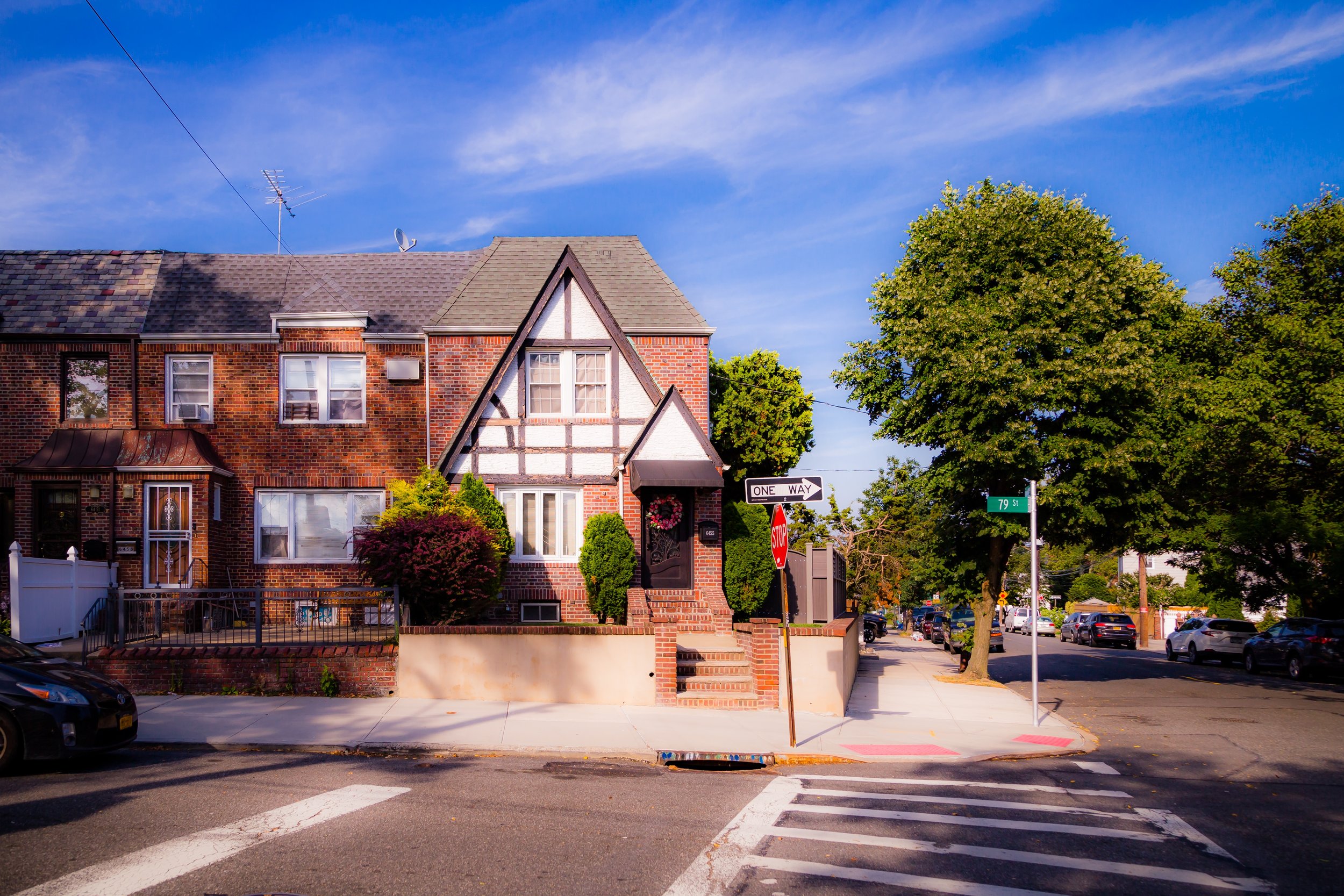 A suburban intersection with a brick house featuring a Tudor-style gable, lush green trees, parked cars, and street signs indicating '79 St' and 'One Way.'