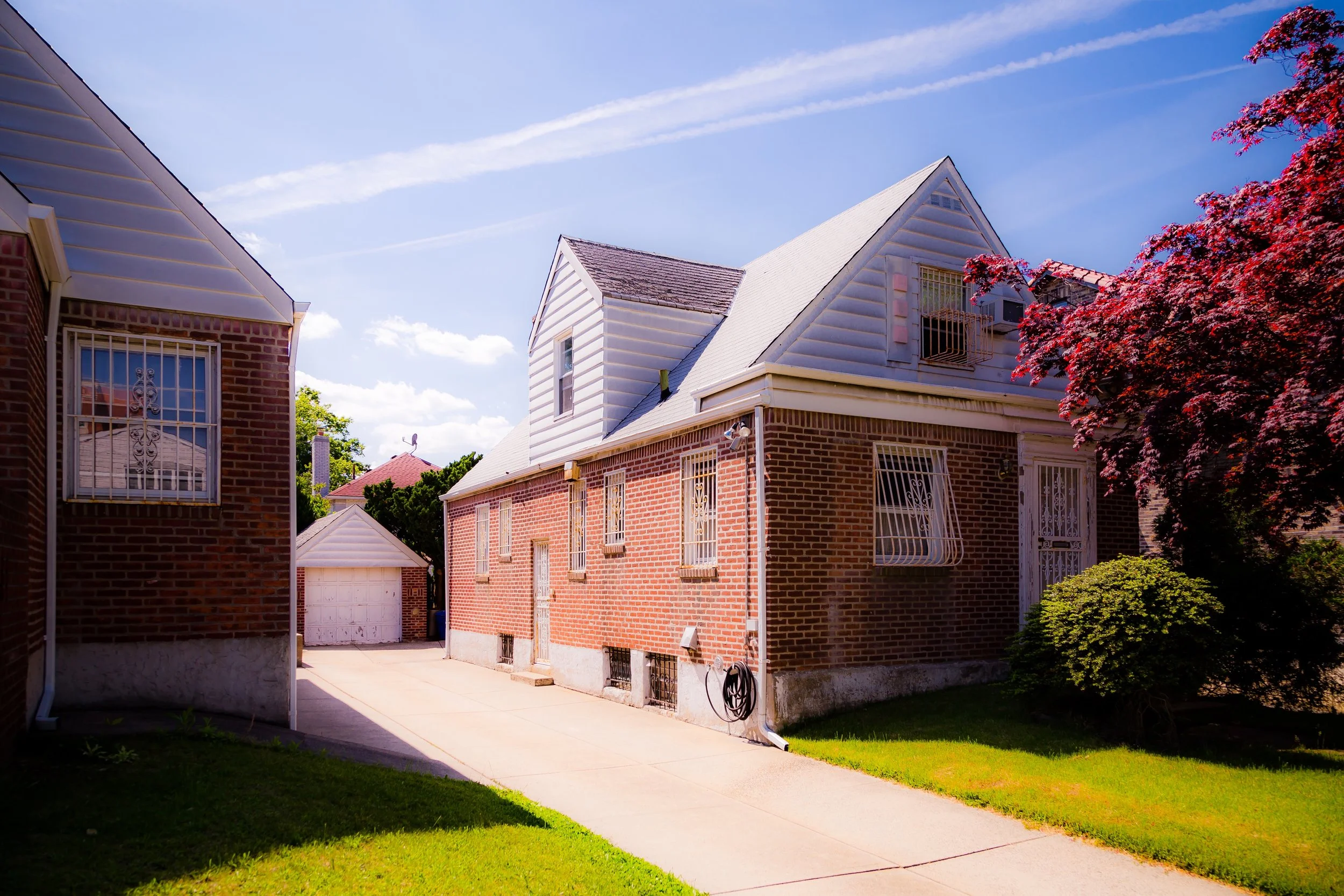 Side view of a brick house with white siding, a concrete driveway, a tree with purple leaves, and a clear blue sky.
