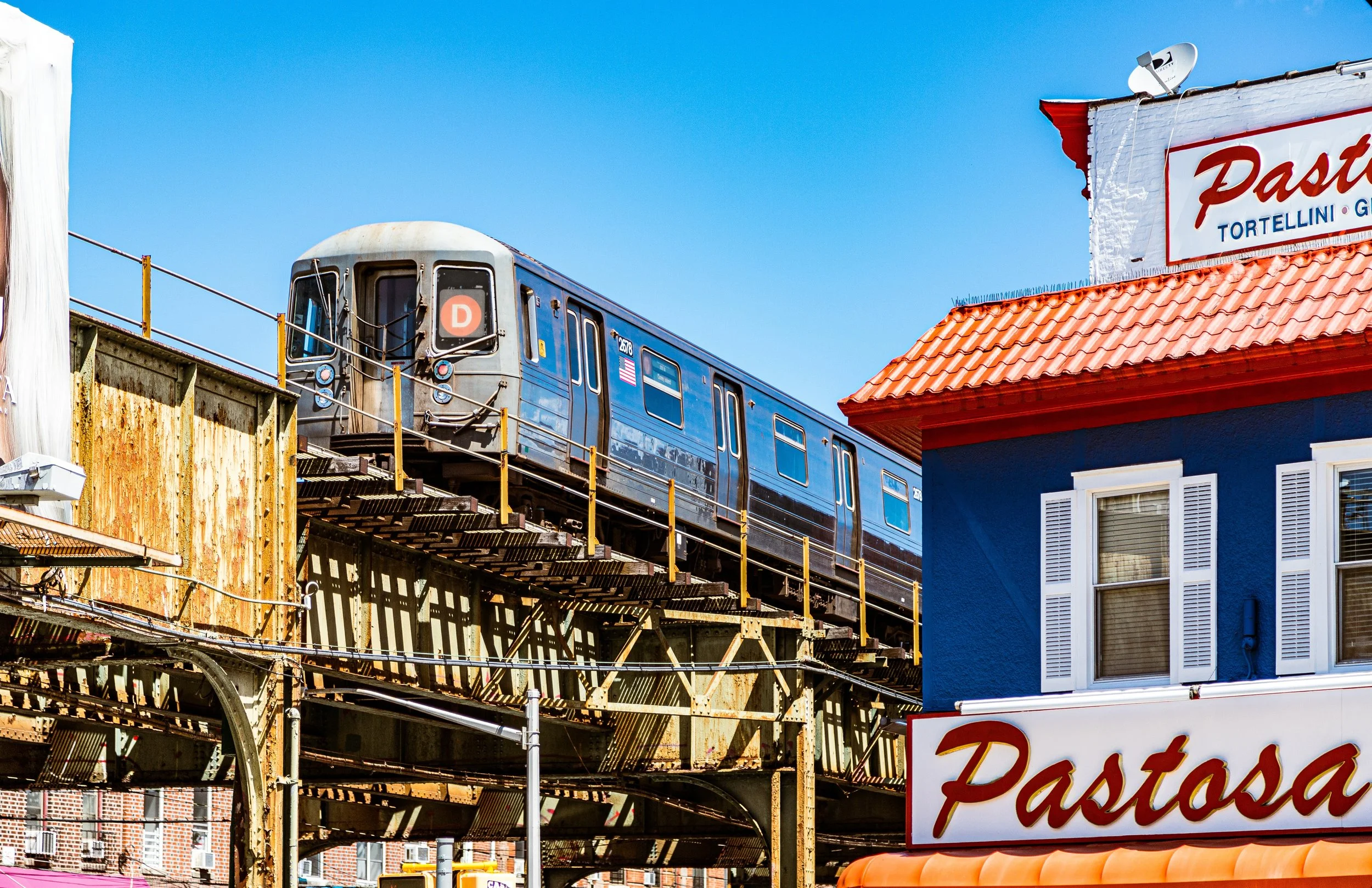 A blue subway train on an elevated track passing above a city street with colorful buildings, including a blue building with white shutters and a sign that reads "Pastova" and lists pasta dishes.
