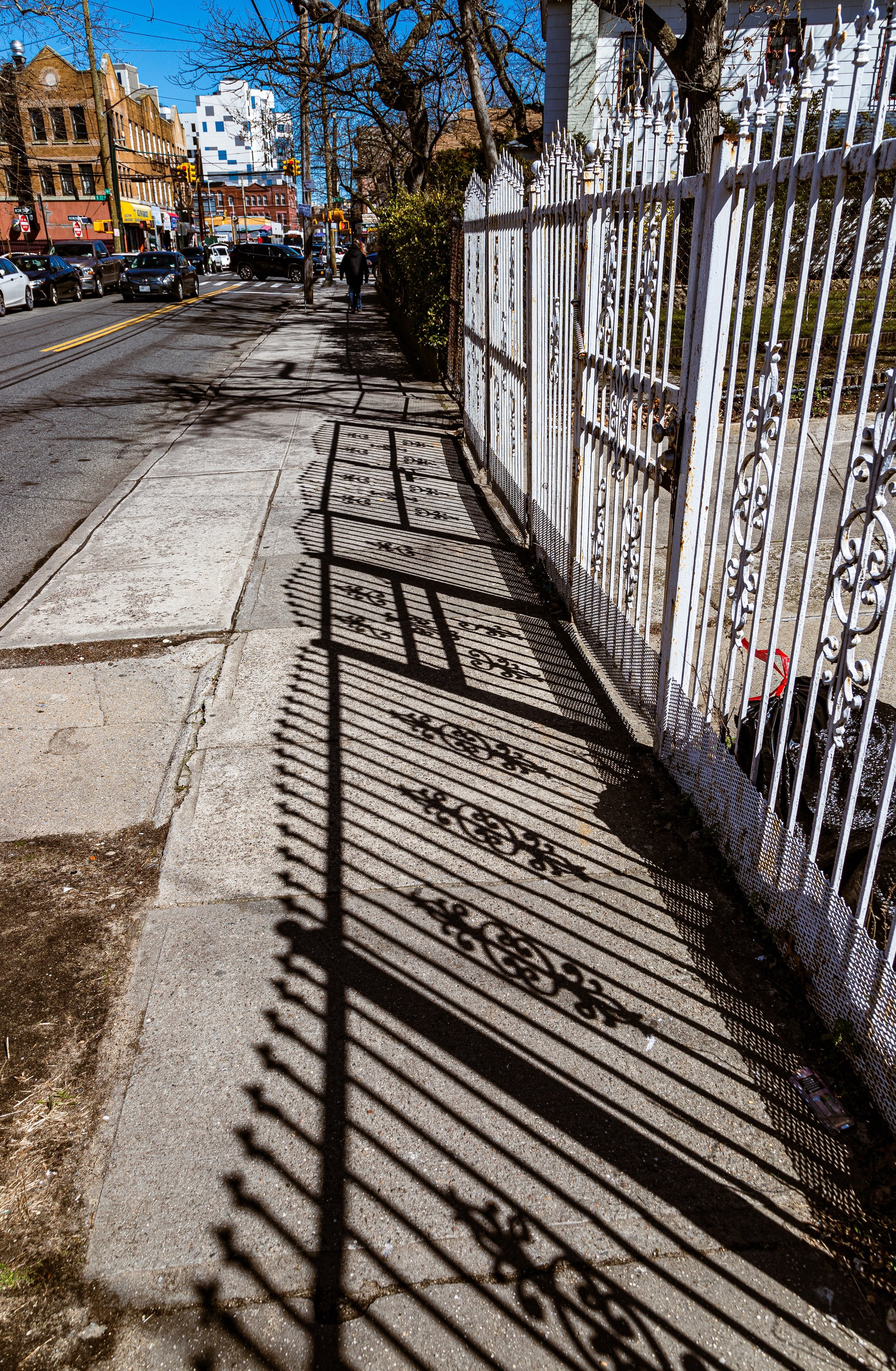 Shadow of an ornate white fence cast on a sidewalk in an urban street scene with parked cars, buildings, and a few pedestrians.