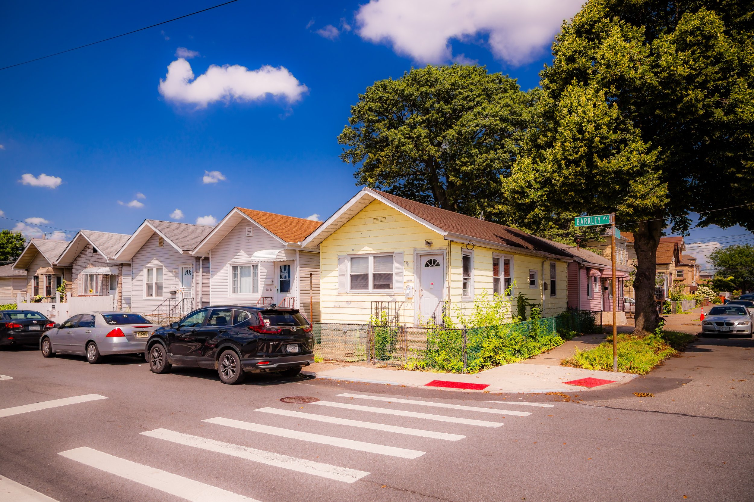 A residential street scene showing a row of small houses with parked cars in front. The houses are pastel-colored, with one yellow house at the corner on Barkley Avenue, under a large leafy tree. The sky is bright blue with a few clouds.