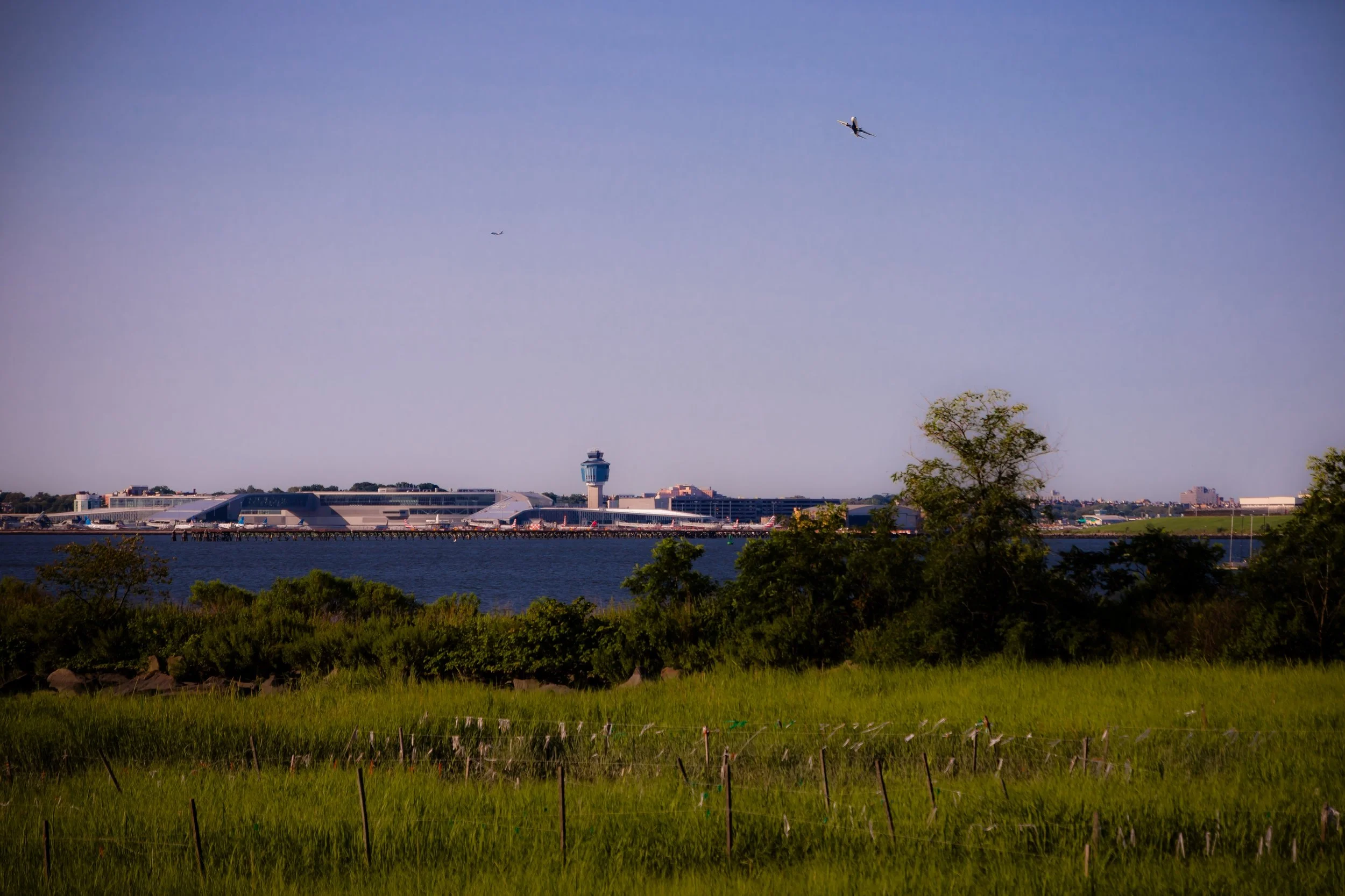 View of an airport terminal across a body of water, with trees and green grass in the foreground, and a airplane flying in the sky.