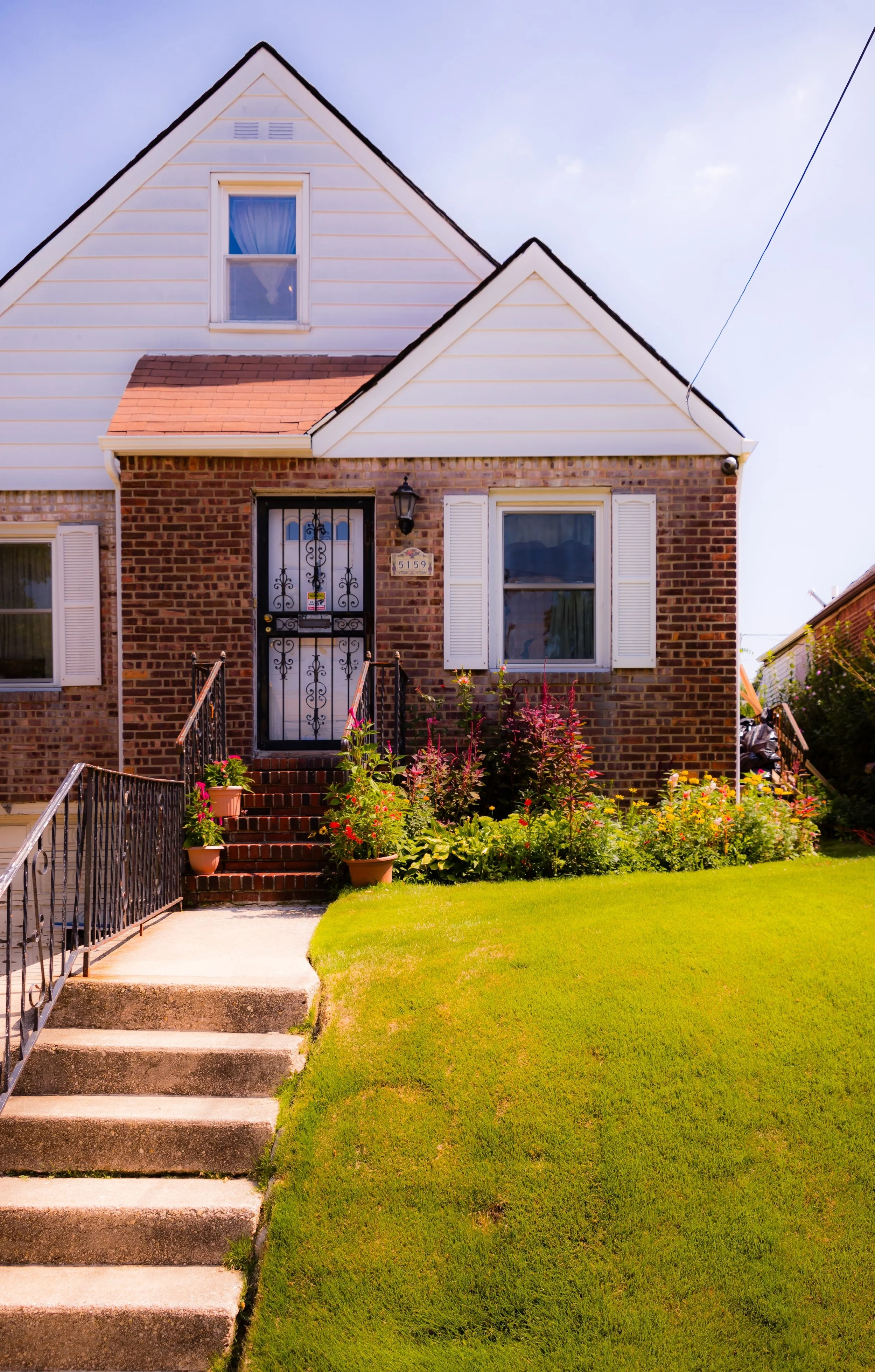 Front view of a two-story house with brick and white siding, featuring a small front garden with potted plants and colorful flowers, concrete steps leading to a black iron door, and white shutters on the windows.