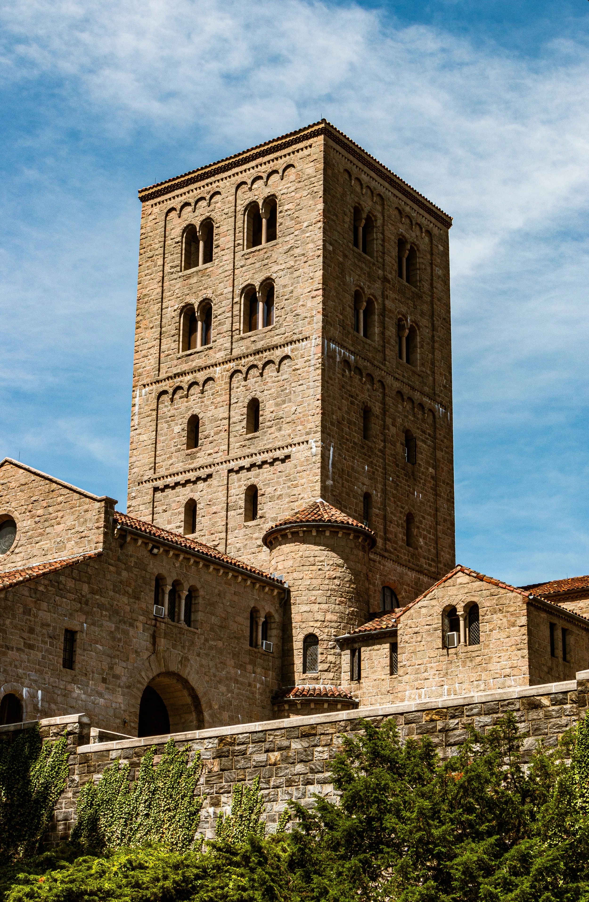 A historic stone tower with arched windows and a castle-like structure, set against a blue sky with scattered clouds.