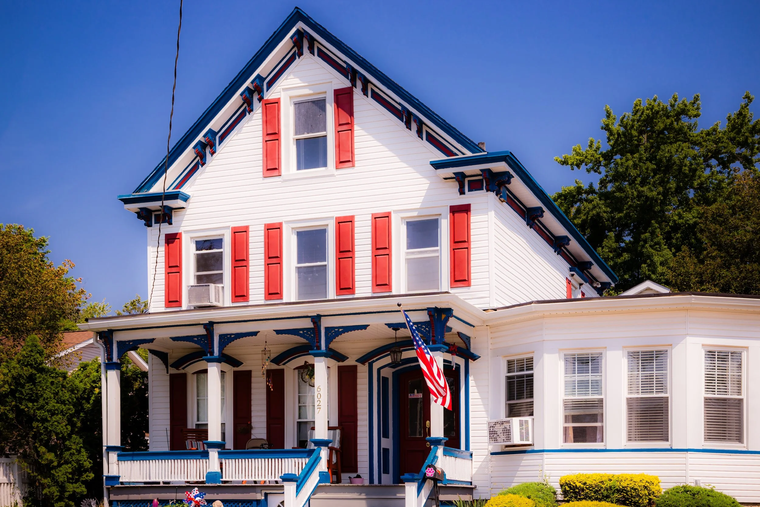 A large, three-story white house with red shutters, blue trim decorative elements, and a porch with columns. An American flag hangs from a pole on the front porch. The house has multiple windows and a gabled roof, with trees and a clear blue sky in the background.
