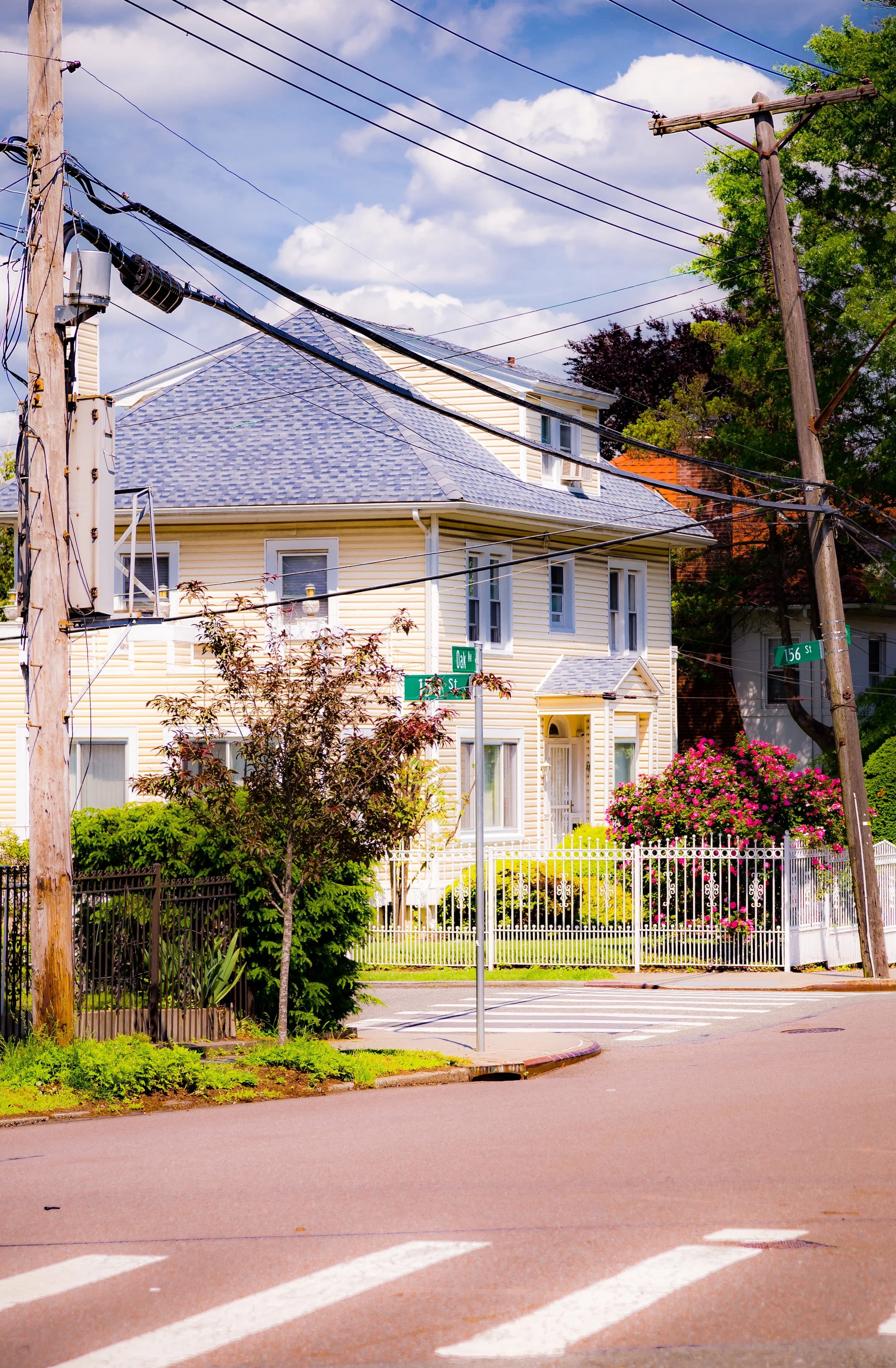 A street corner with a crosswalk, parking signs, and a large yellow house with a white fence and pink flowering bushes, under a blue sky with clouds.