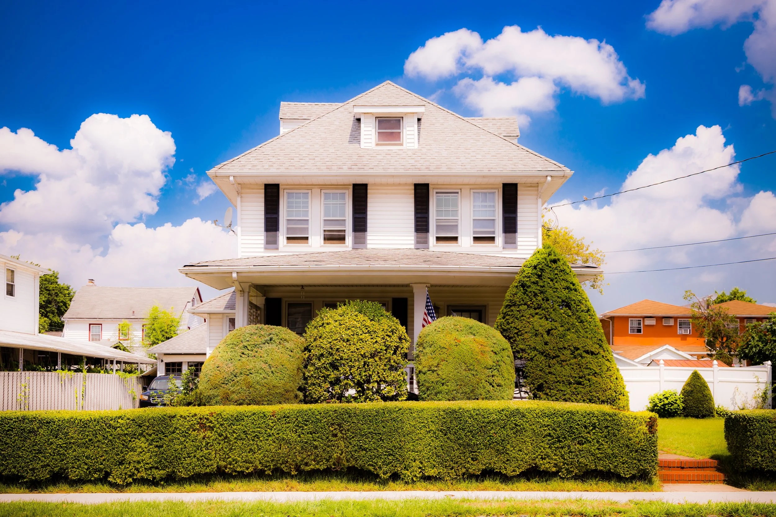 A white, two-story house with a gabled roof, several windows, and black shutters. The front yard is landscaped with neatly trimmed bushes and hedges under a bright blue sky with white clouds.