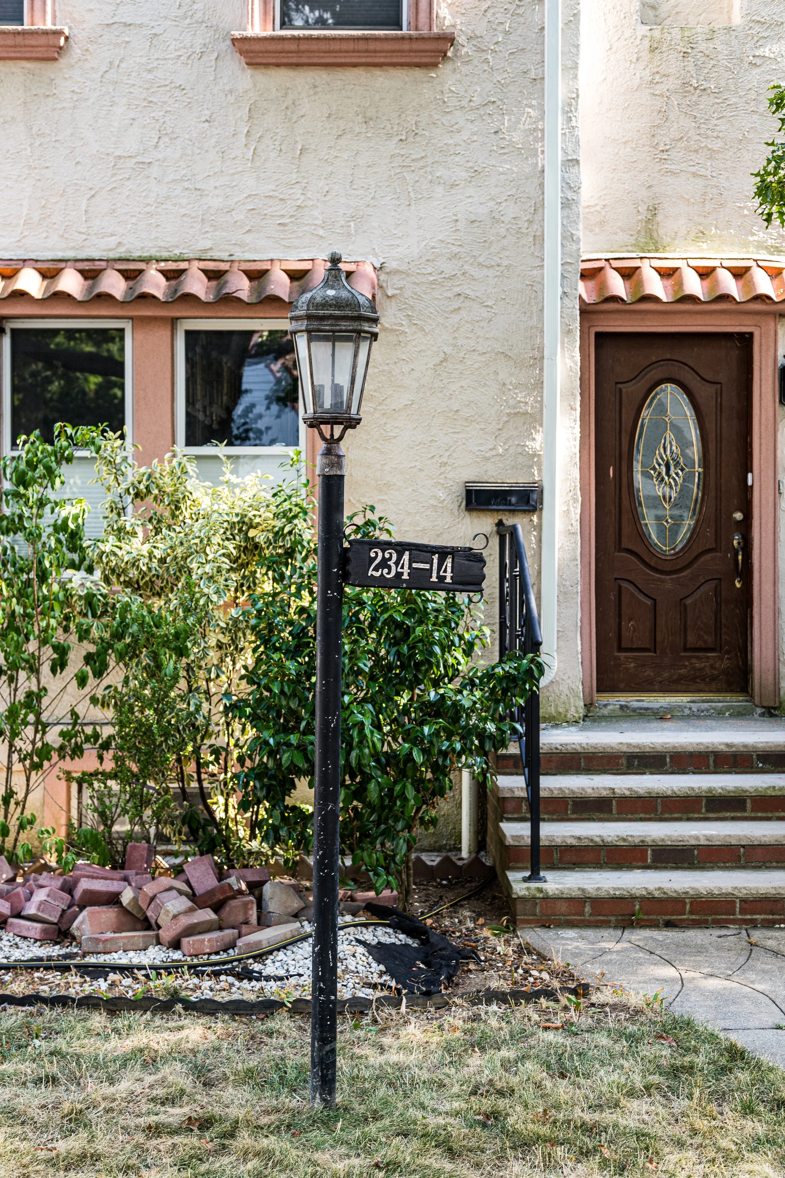 A house with a wooden front door, concrete steps, a black mailbox, a black vintage-style lamp post with a house number sign, and lush green bushes in the garden.