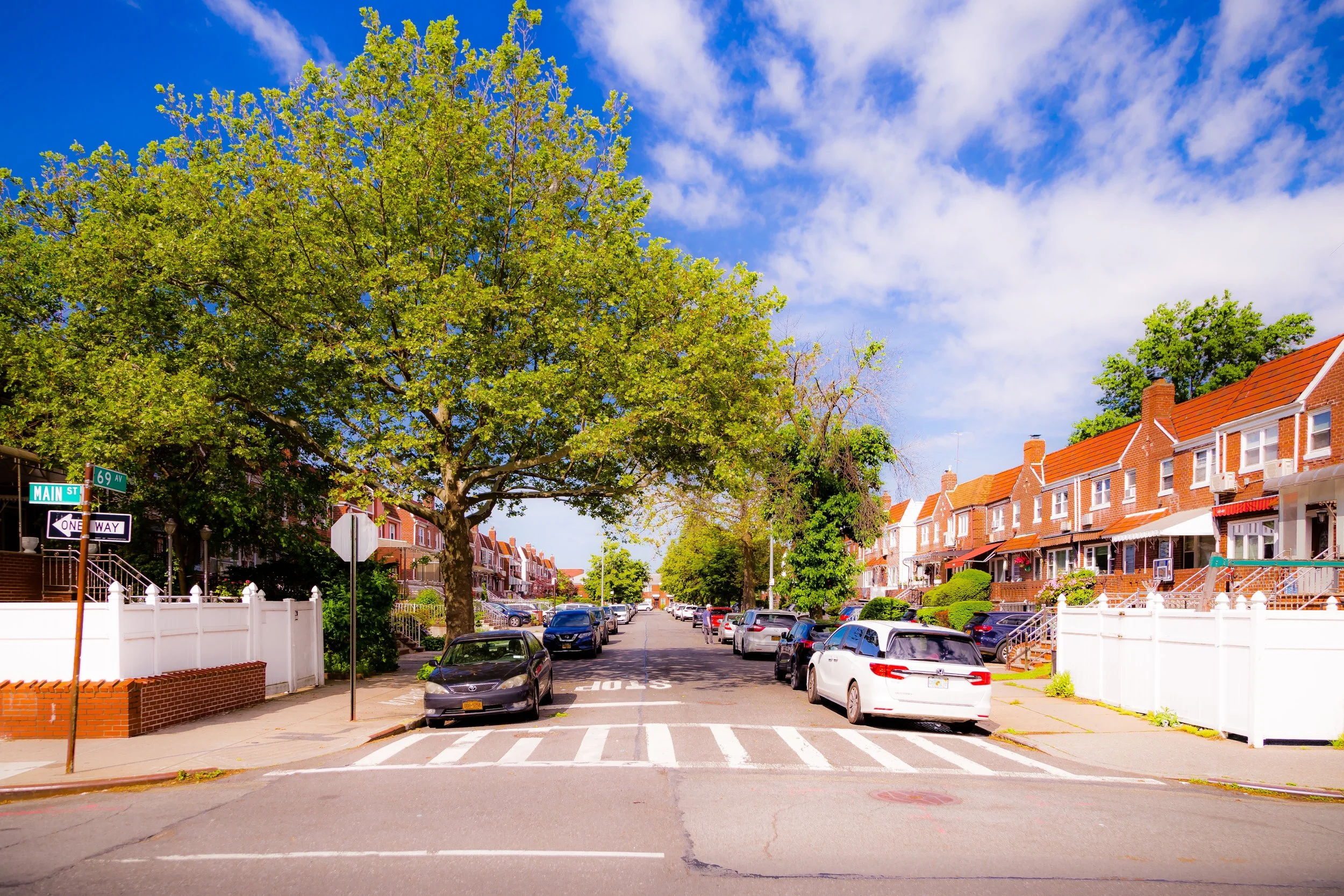 Residential street with cars parked along the curb, large trees and row houses with red roofs, blue sky with some clouds.