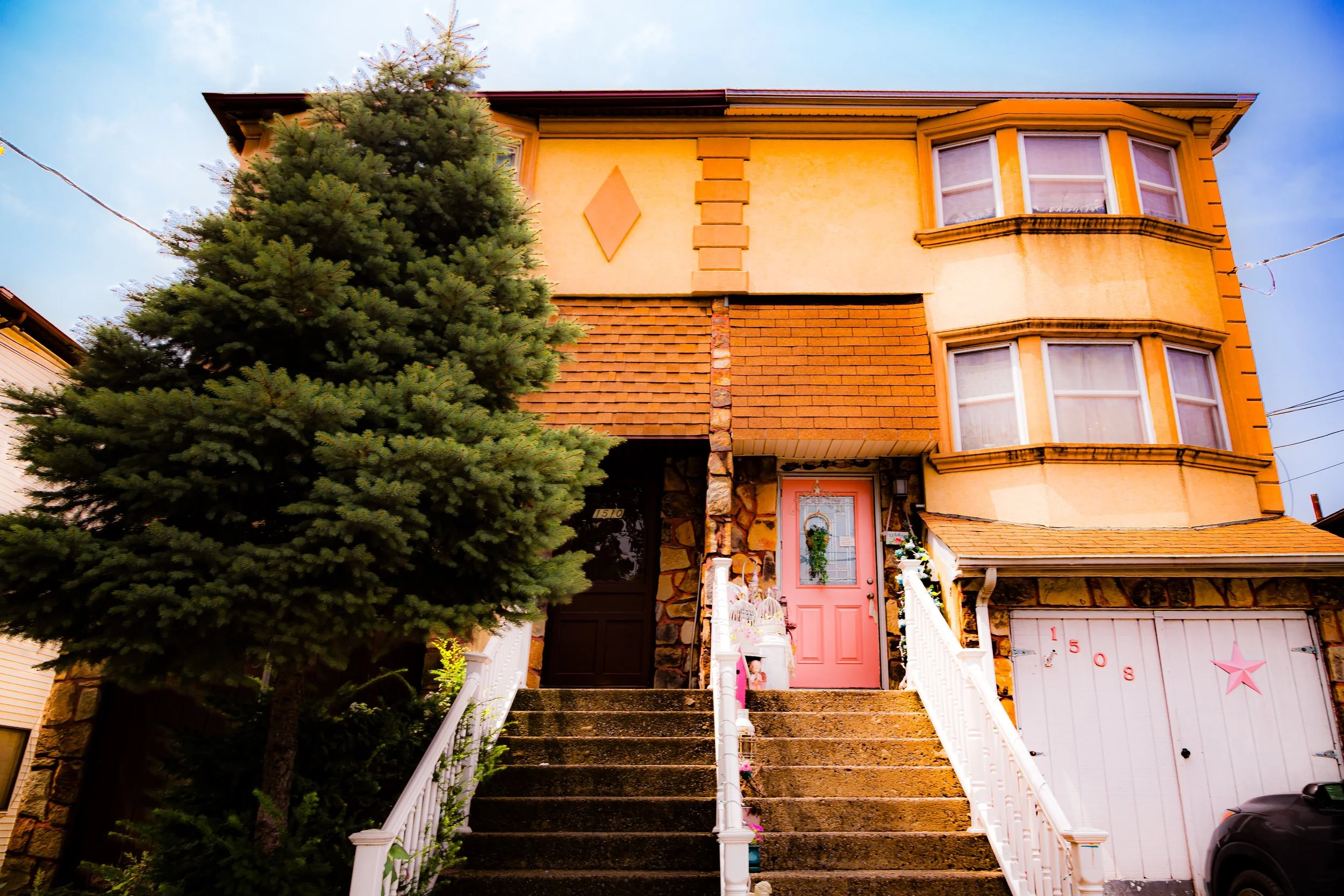 A multi-story house with a pink front door, a large evergreen tree on the left, and stairs with white railings leading up to the entrance. The house has a yellow exterior with decorative brick and stone accents.
