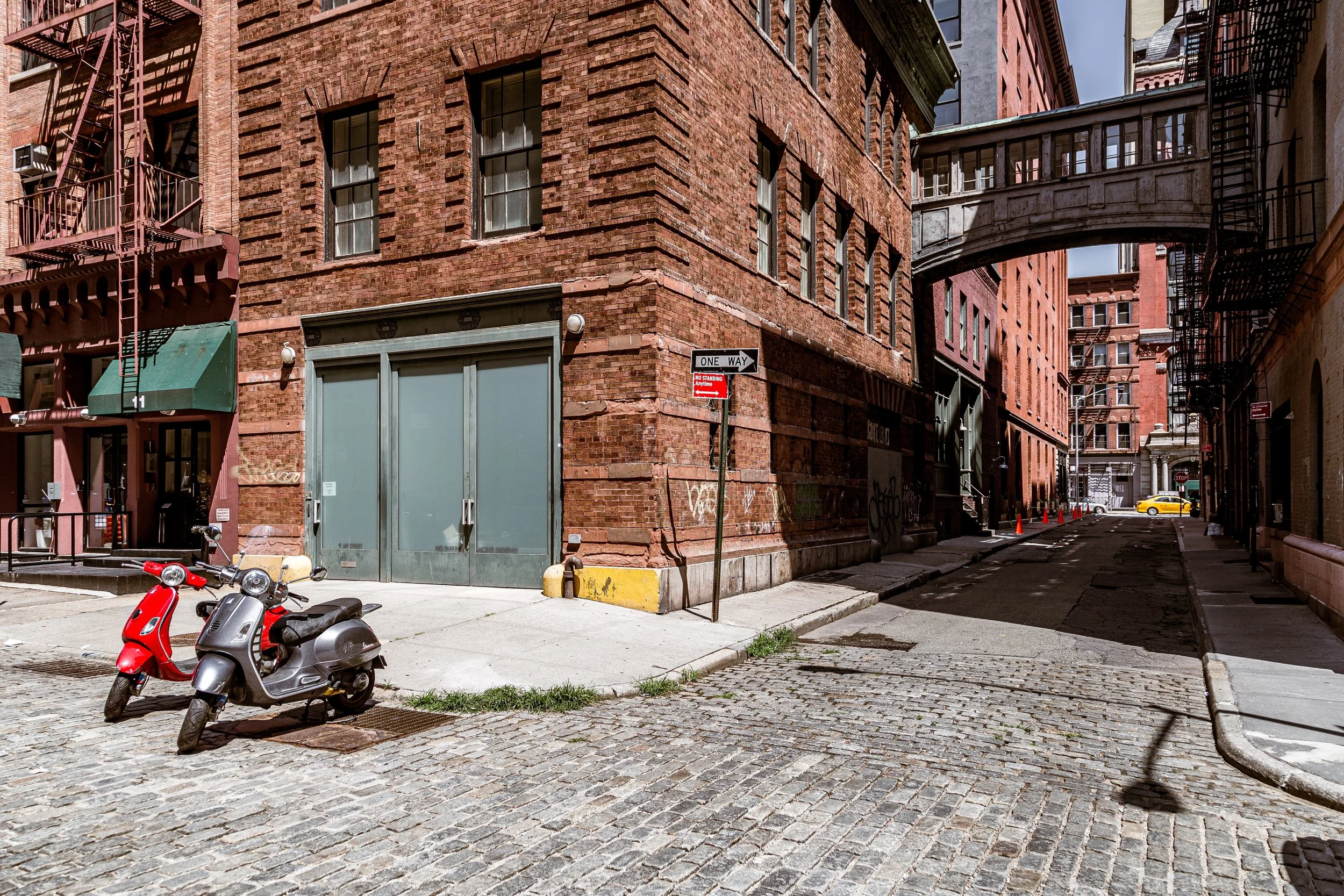 Street view with cobblestone pavement, red-brick buildings, a small scooter parked on the sidewalk, a metal fire escape on the building, a bridge connecting two buildings, and a yellow taxi in the distance.