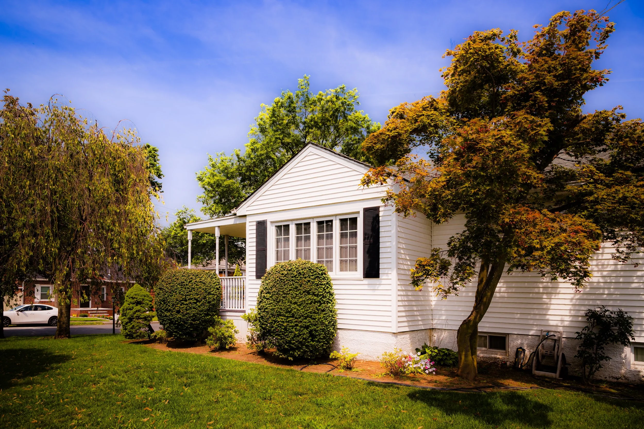A white house with black shutters, a porch, and a pitched roof, surrounded by green trees, bushes, and a well-maintained lawn on a sunny day.