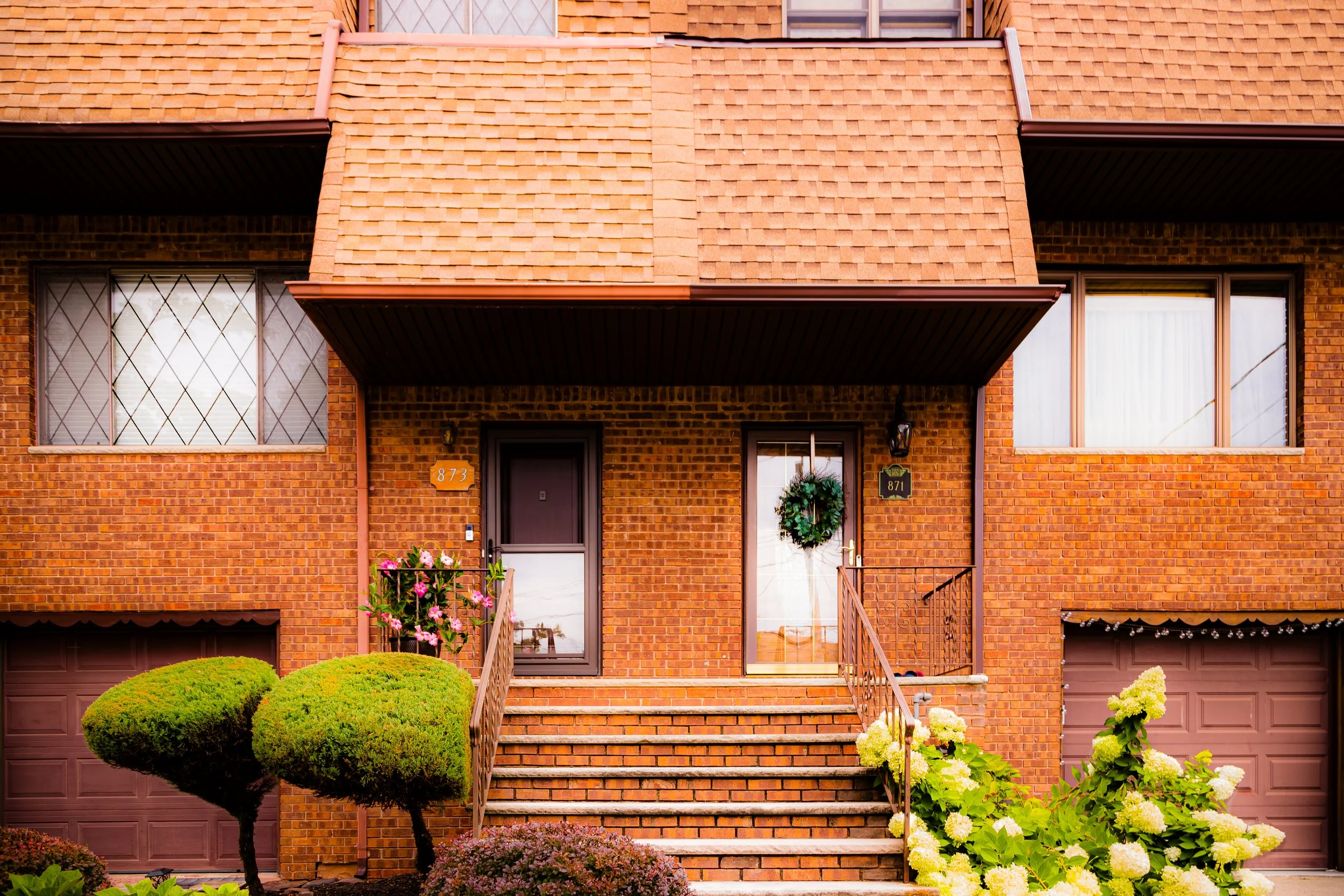 Brick residential building with stairs leading to front doors, decorated with a wreath and flowers, with two windows and garage doors.