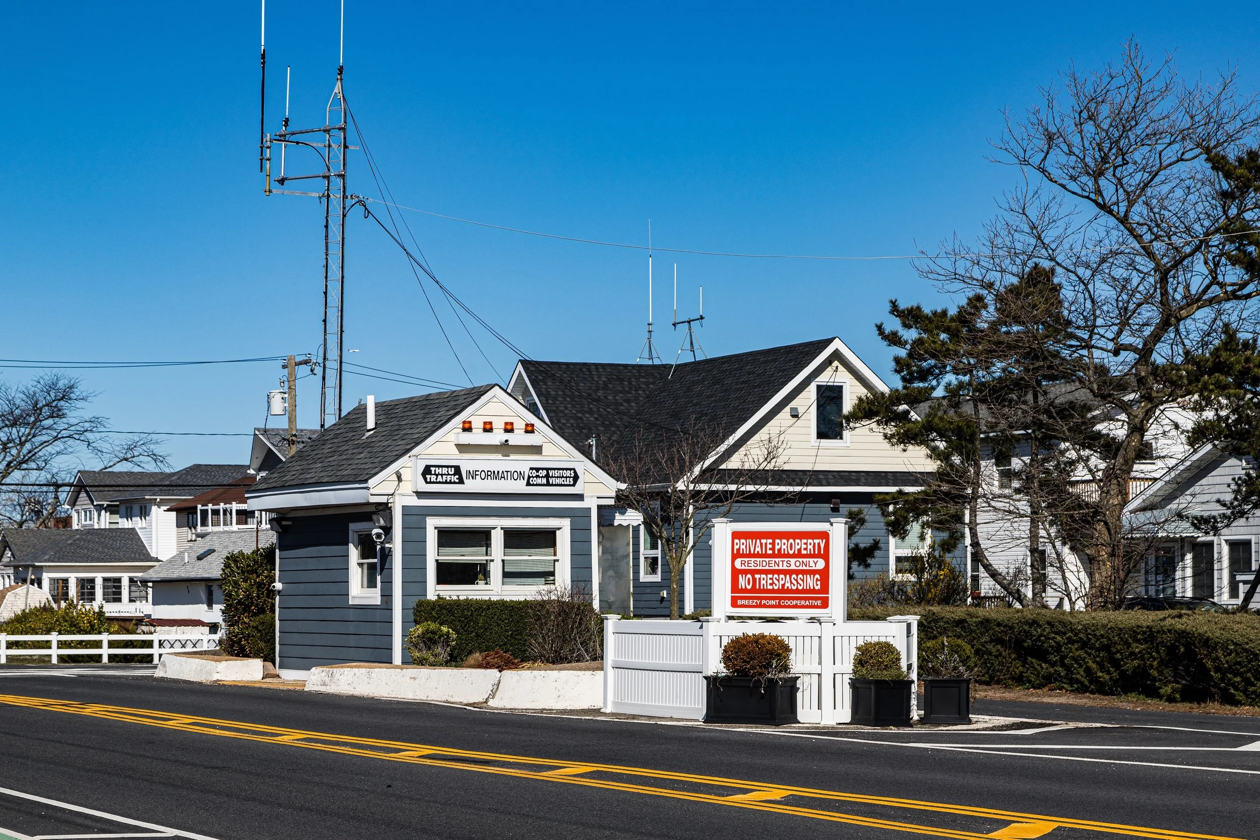 A small blue and white building on the side of a road with a sign that says 'Thru Traffic Information'. There is a white fence with potted plants and a red and white sign that reads 'Private Property, Residents Only, No Trespassing'. Several houses and trees are in the background under a clear blue sky.