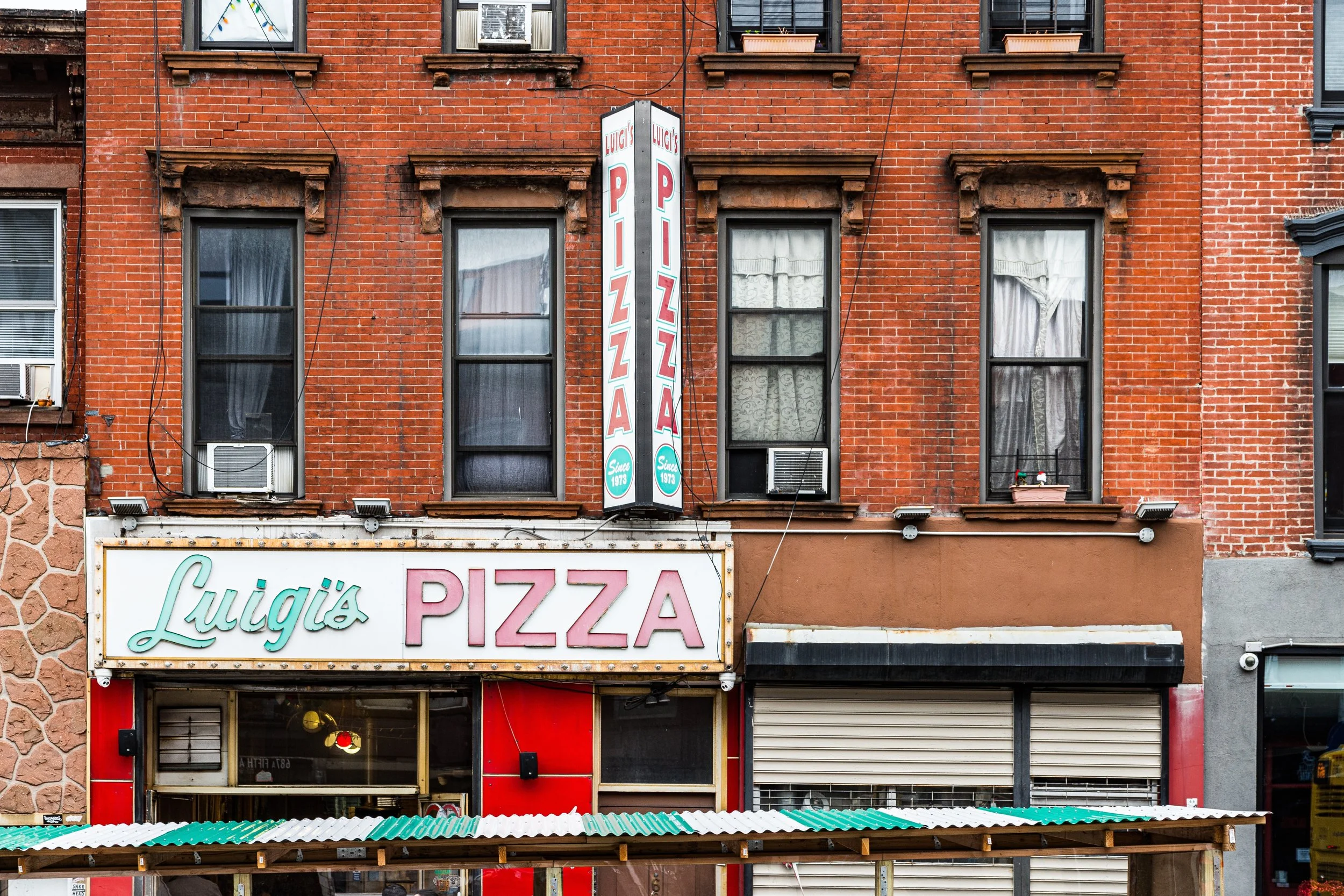 A brick building with a pizza restaurant named Luigi's Pizza. The sign displays the restaurant's name and a vertical banner with the same name. The windows have curtains and air conditioning units, and there is a small flowerpot on one window sill.