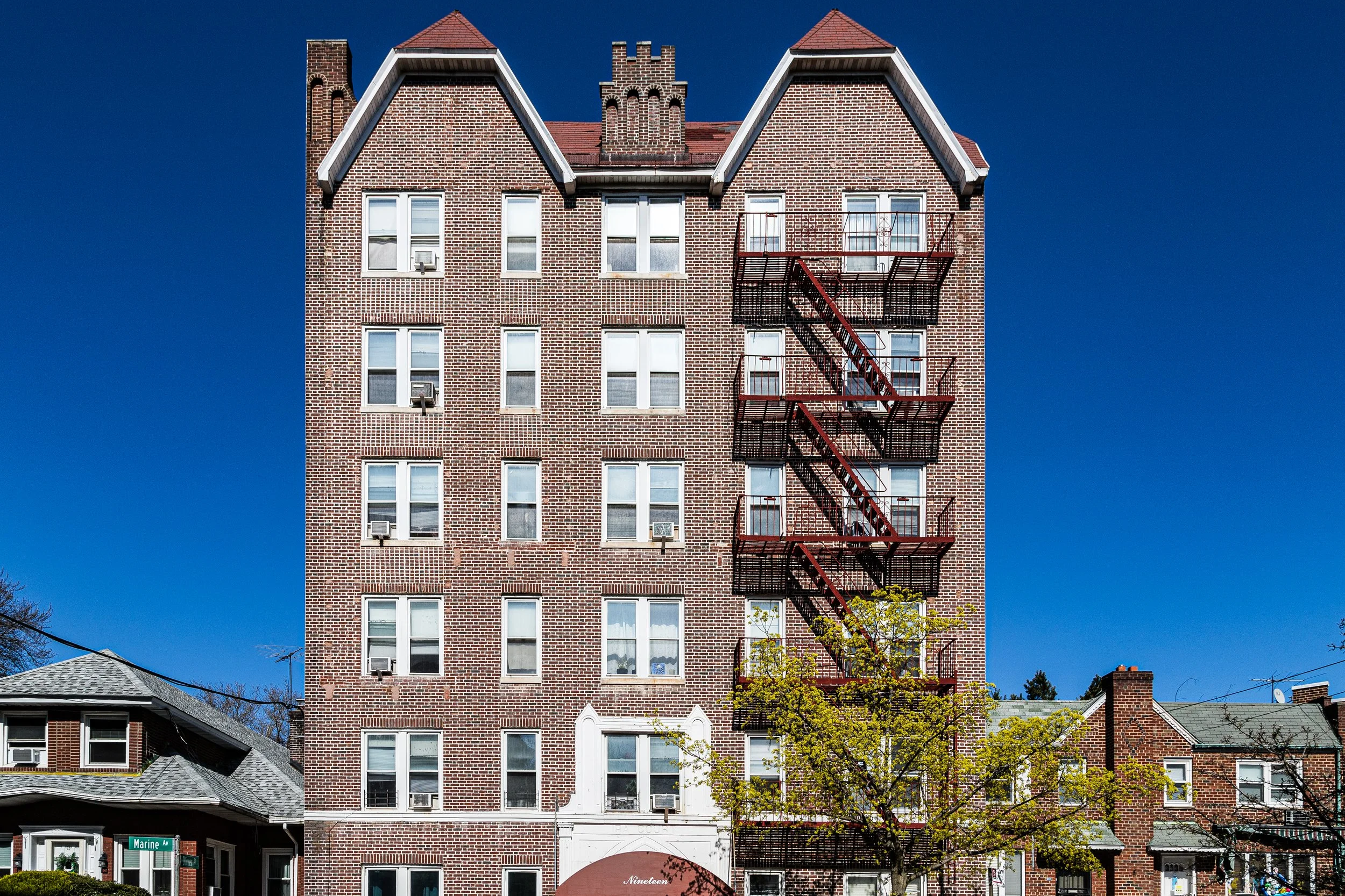 A multi-story brick apartment building with fire escape stairs on the right side, blue sky background, and a small tree with green leaves in front.