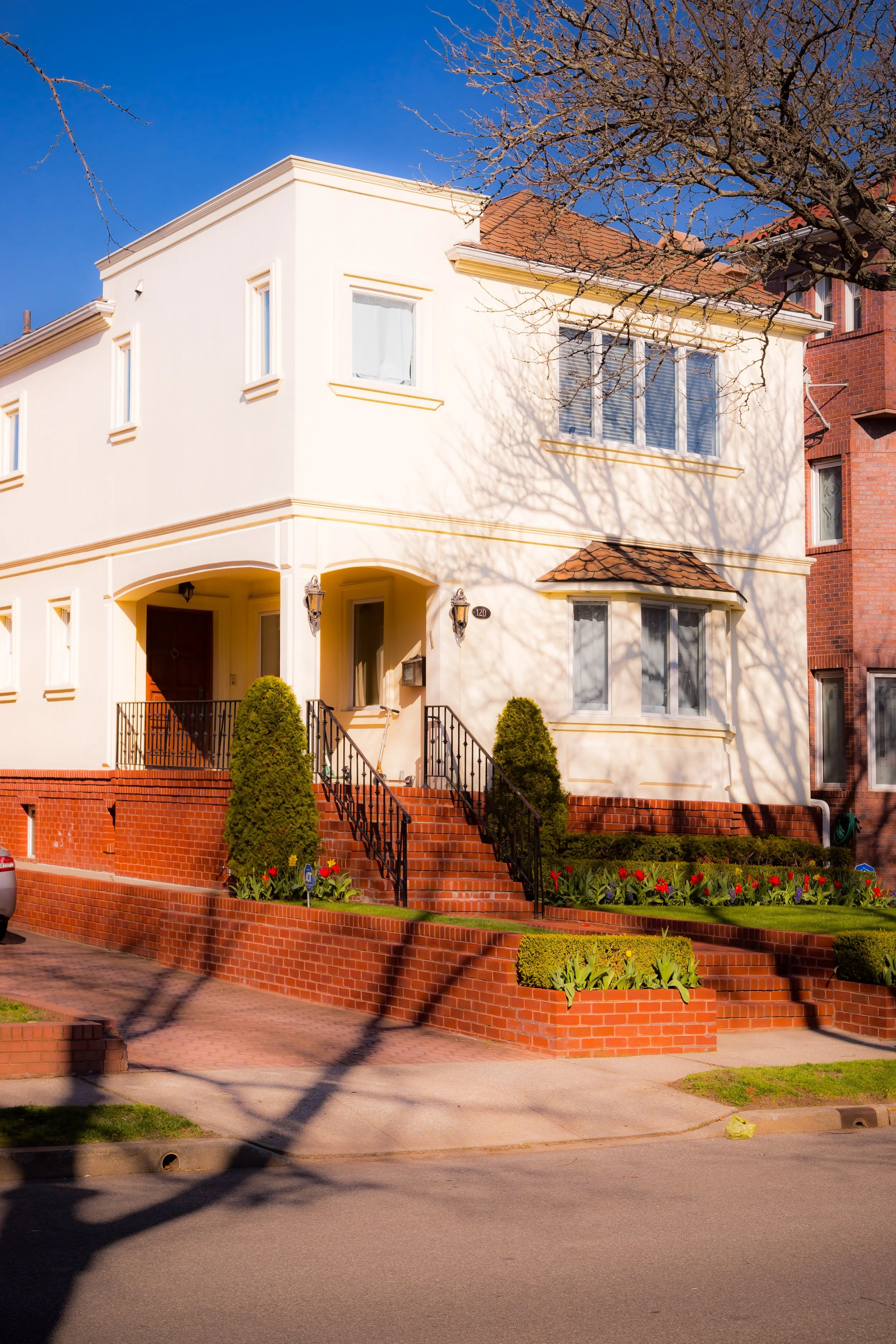 A two-story white house with brick steps and red brick foundation, black metal railings, and a well-maintained garden with bushes and flowers in front. Some bare tree branches cast shadows on the house, and the sky above is clear and blue.