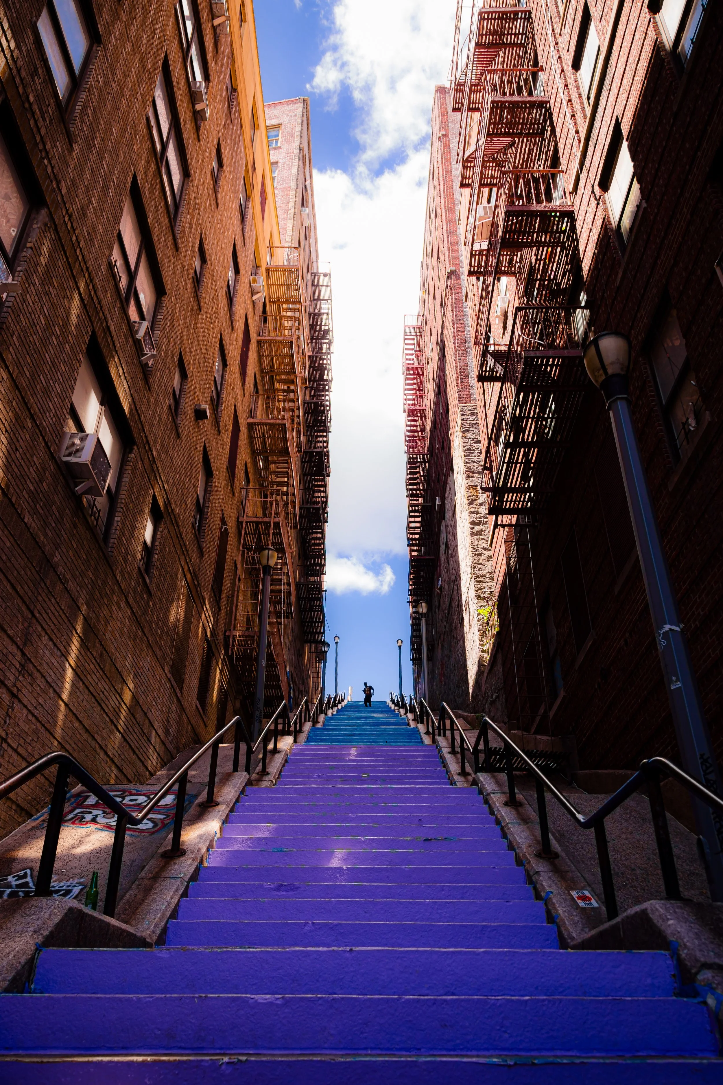 A long outdoor staircase painted purple, flanked by black railings, ascending between two tall brick apartment buildings with fire escapes on a city street. Two people are near the top of the stairs, under a partly cloudy sky.