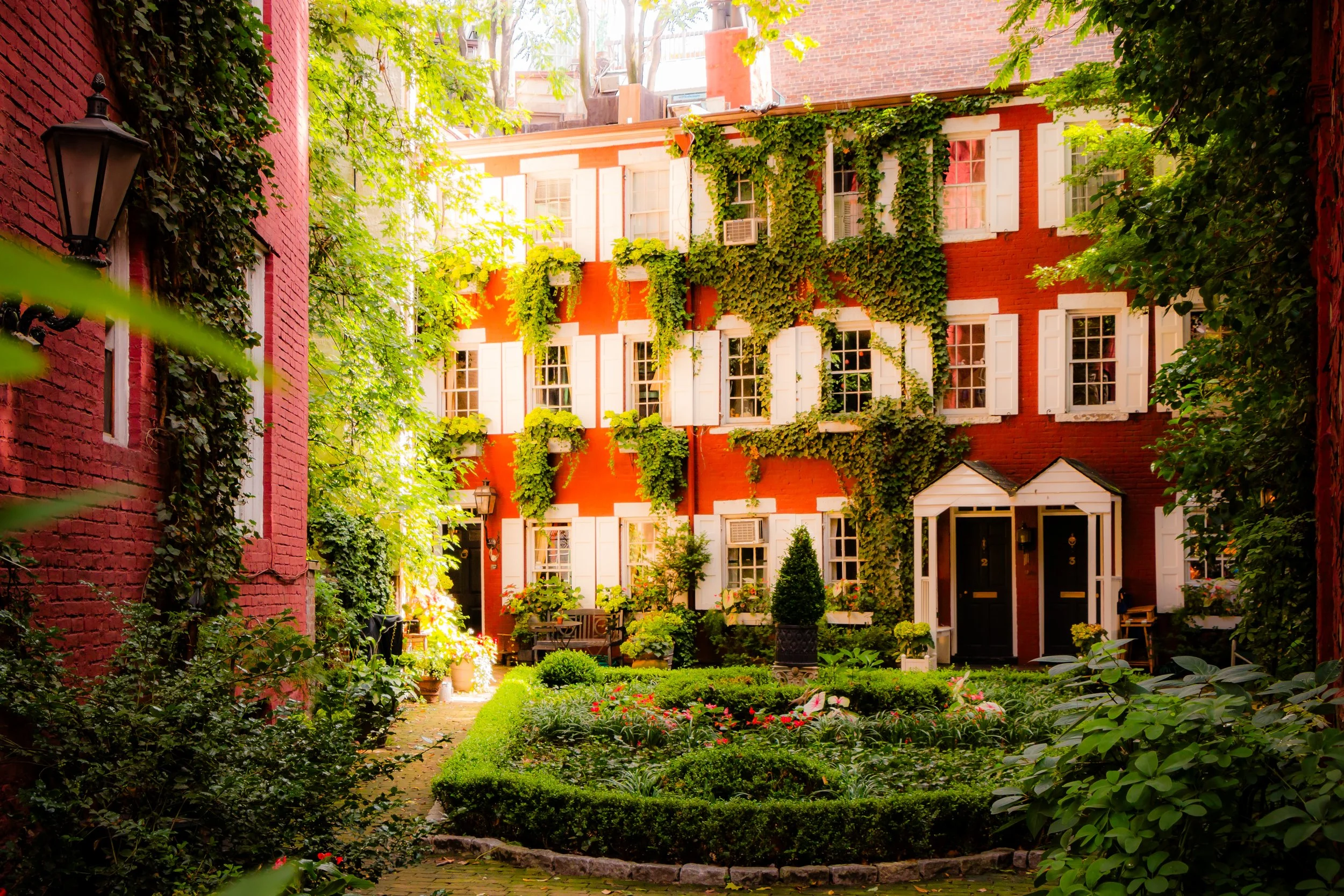 A lush courtyard garden with a red brick building covered in green ivy, white window shutters, and a pathway through well-maintained flower beds.