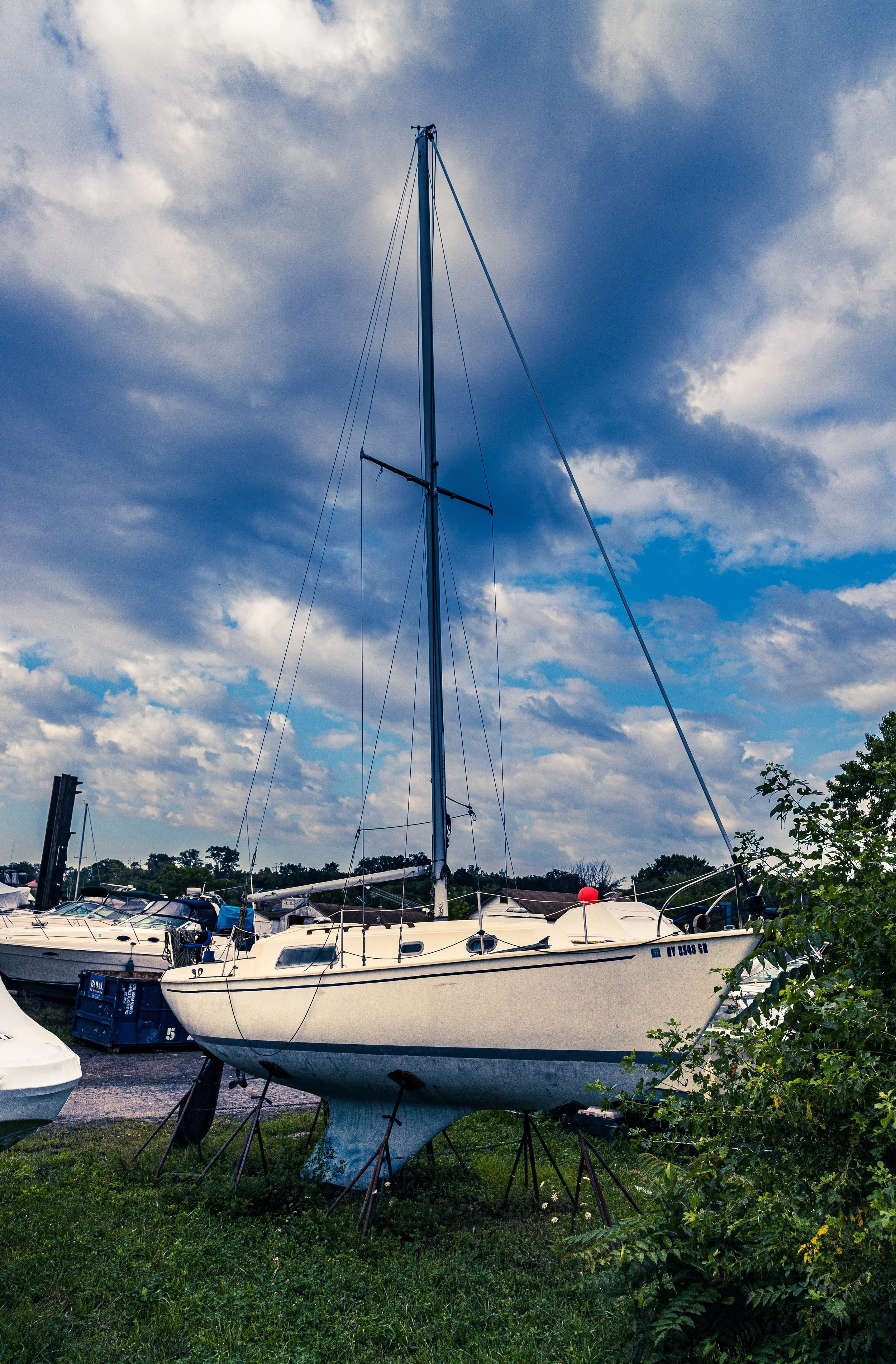 A sailboat on land with a cloudy sky in the background, surrounded by other boats at a marina.