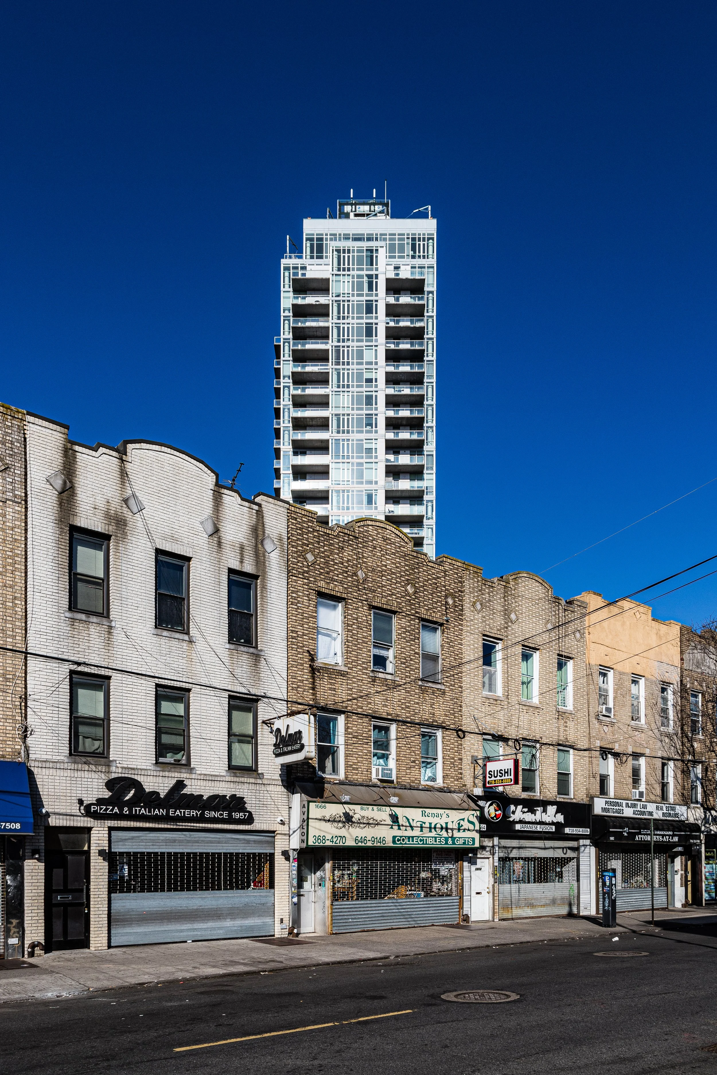 City street with older brick buildings and shops in the foreground, and a tall modern glass skyscraper in the background against a clear blue sky.
