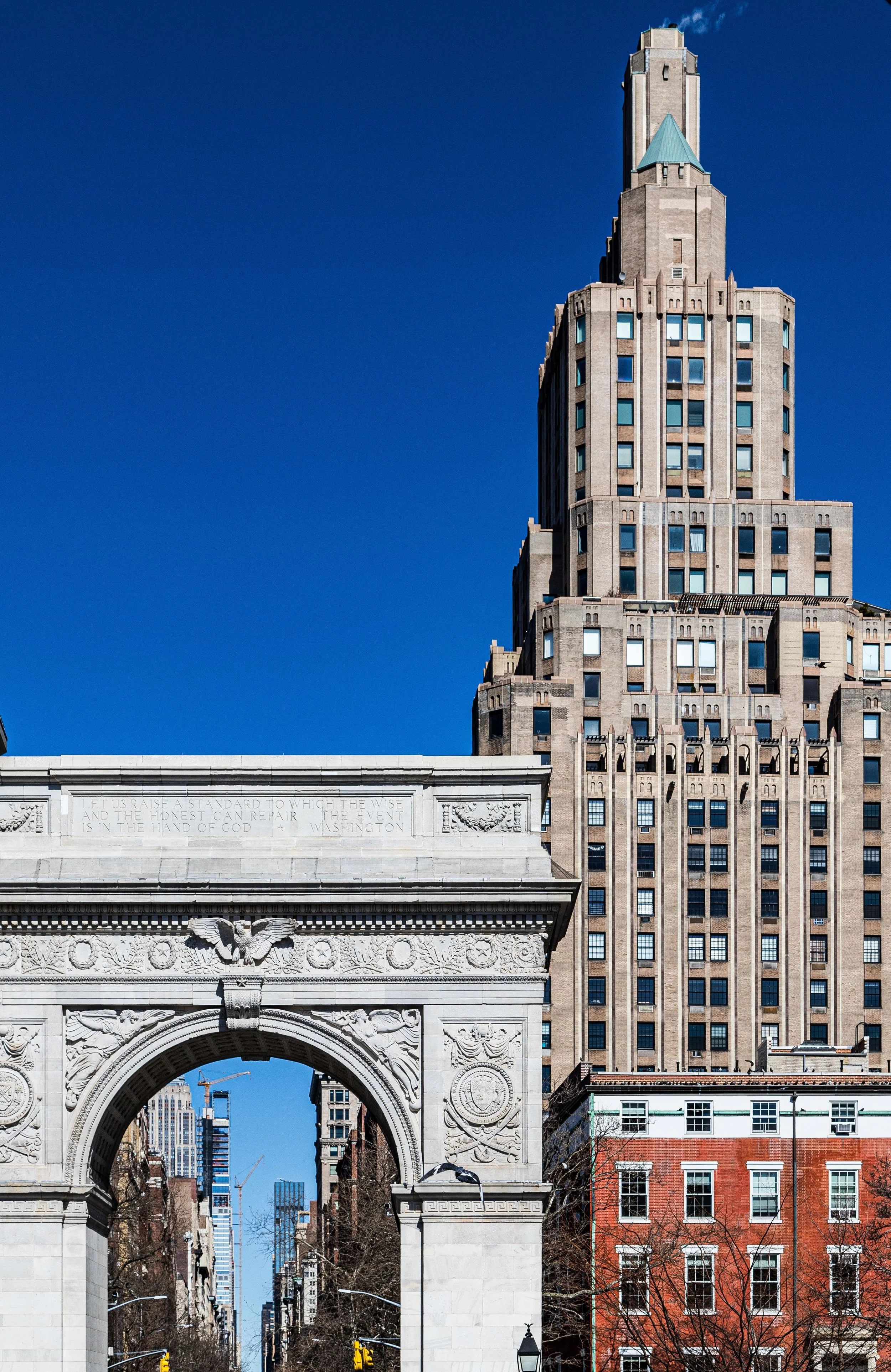 View of a historic white stone arch with an eagle sculpture, ornate carvings, and a quote, with a cityscape featuring tall buildings and a clear blue sky in the background.