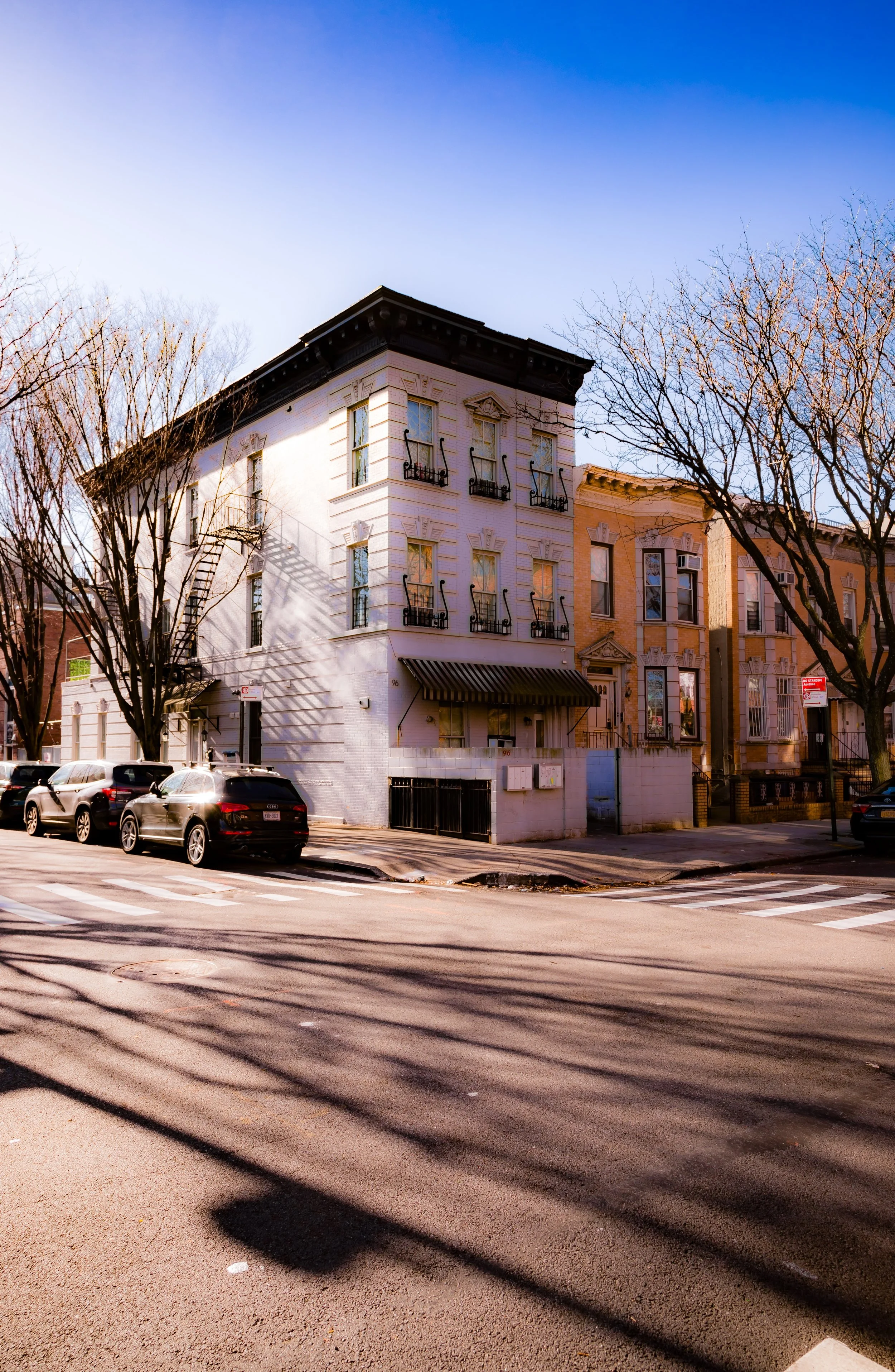 A city street corner with a white three-story building, parked cars, leafless trees, and a clear blue sky.