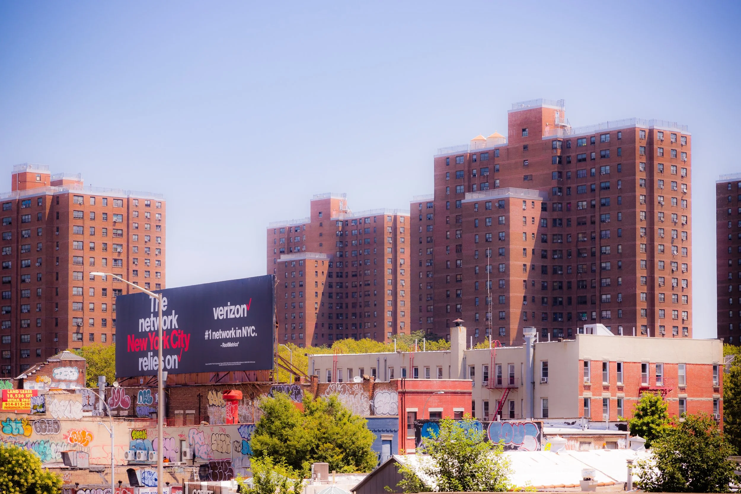 Cityscape featuring tall brown brick apartment buildings, a billboard advertising Verizon and NYC network, graffiti-covered buildings, trees, and clear blue sky.