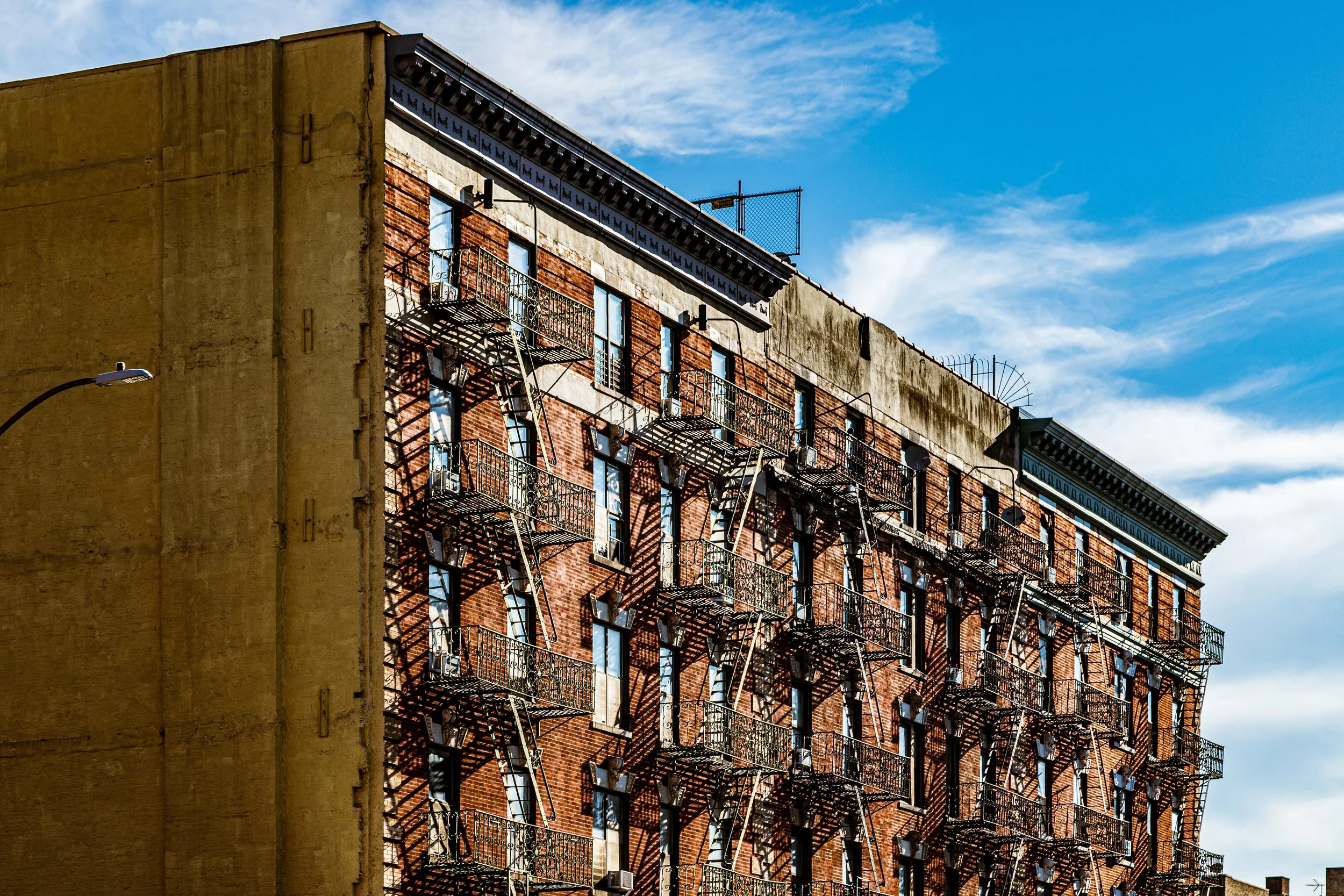 A multi-story brick building with fire escapes on the front, against a partly cloudy blue sky.