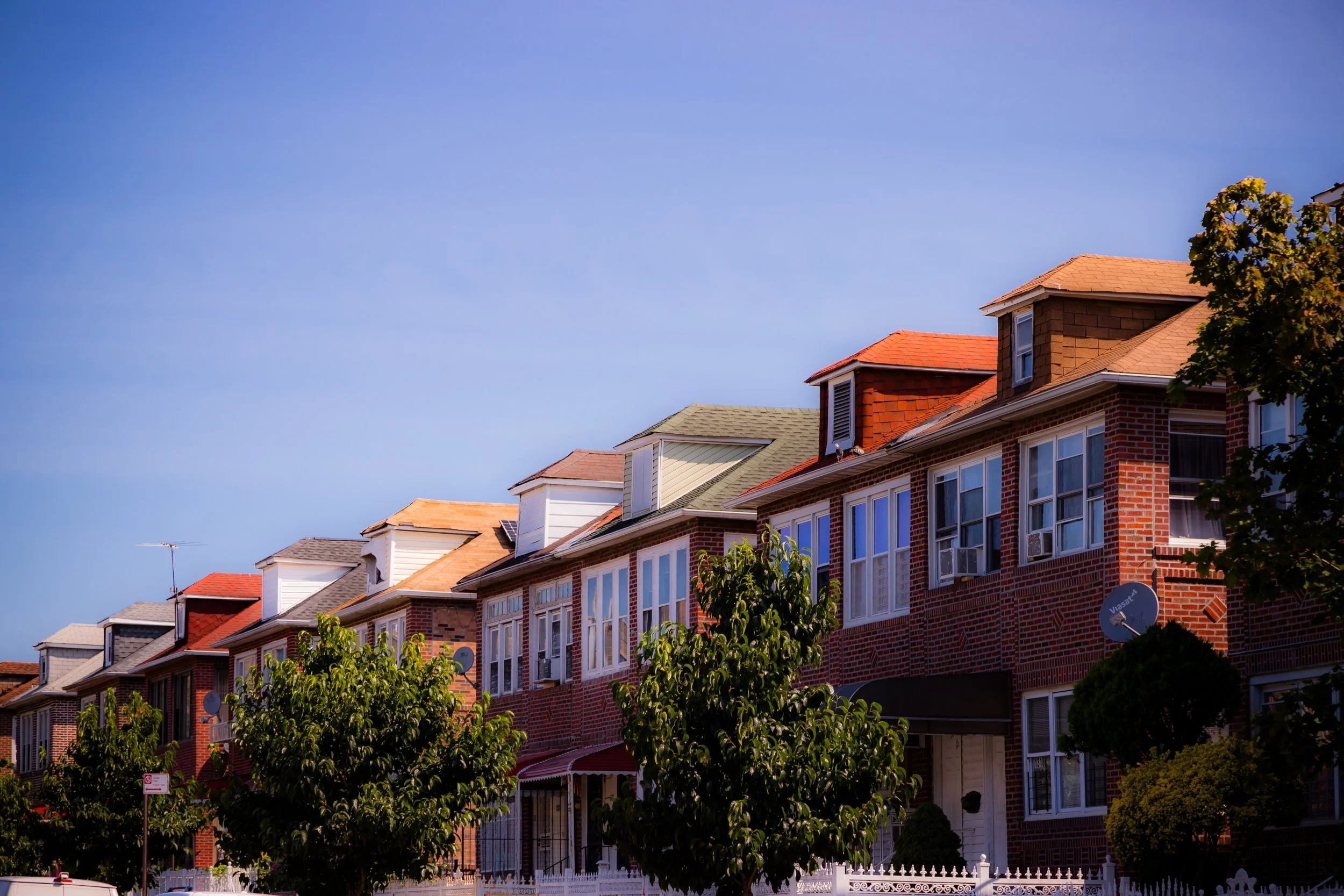 A row of brick houses with various colored roofs and trees in front, under a clear blue sky.