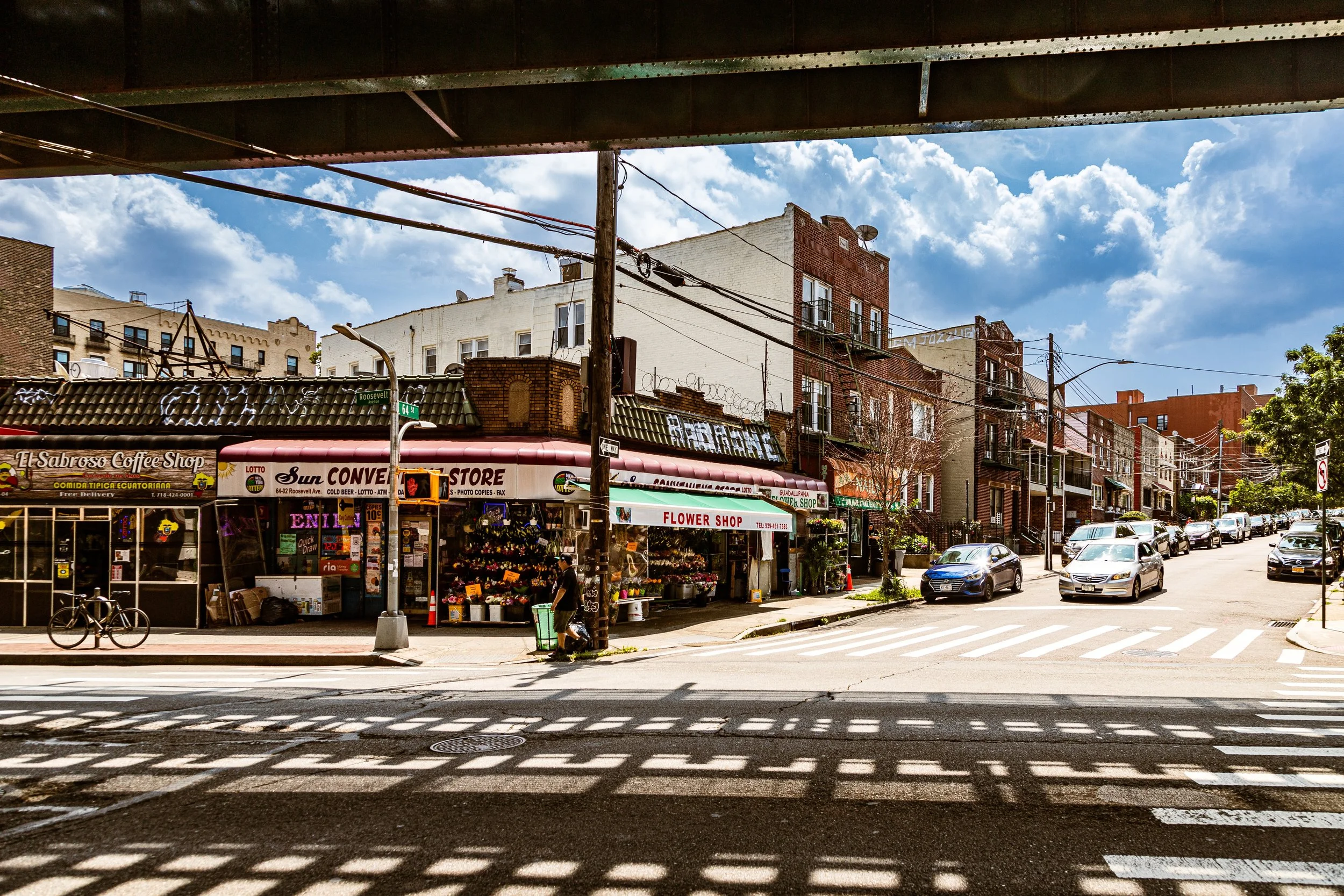 Urban street scene viewed from under a bridge, with storefronts, parked cars, and a clear blue sky.