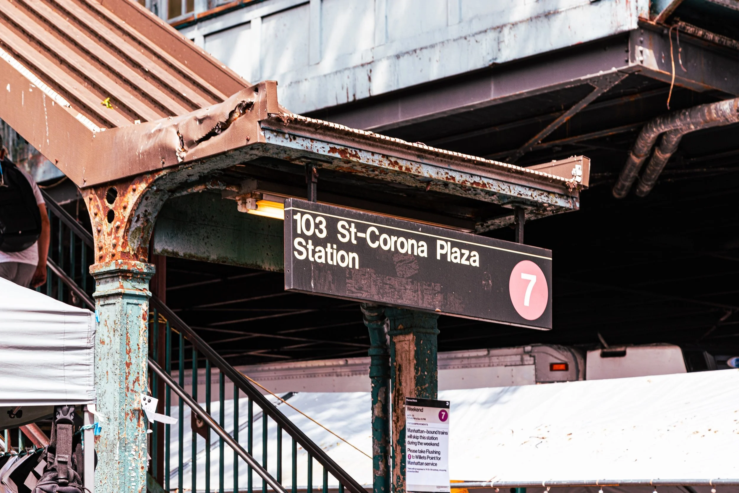 A subway station sign reading '103 St-Corona Plaza Station' with a pink circle containing the number 7, under a rusted metal canopy, with some snow and part of a person visible.