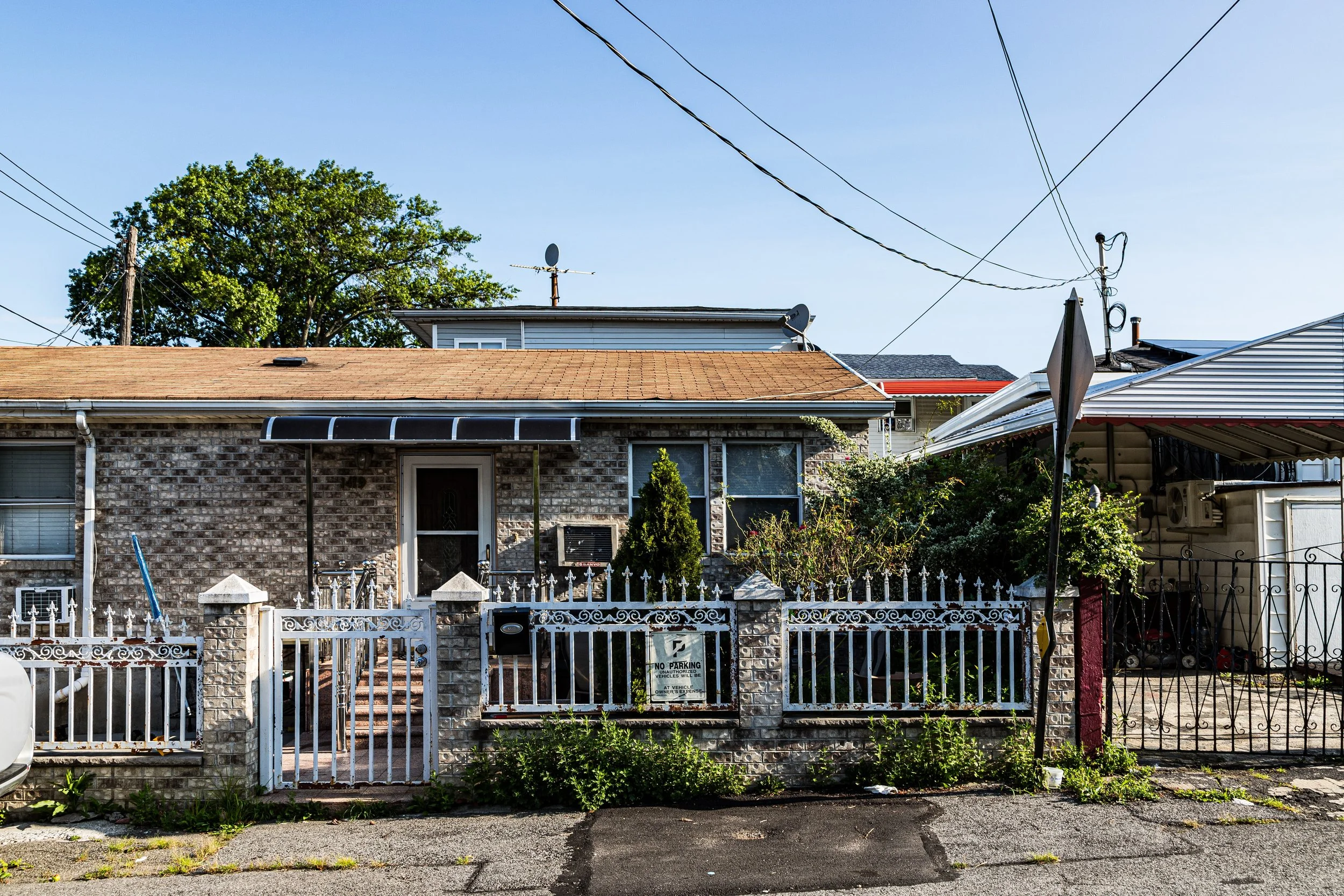 Front view of a small brick house with a white fence and gate, surrounded by greenery, in an urban neighborhood.
