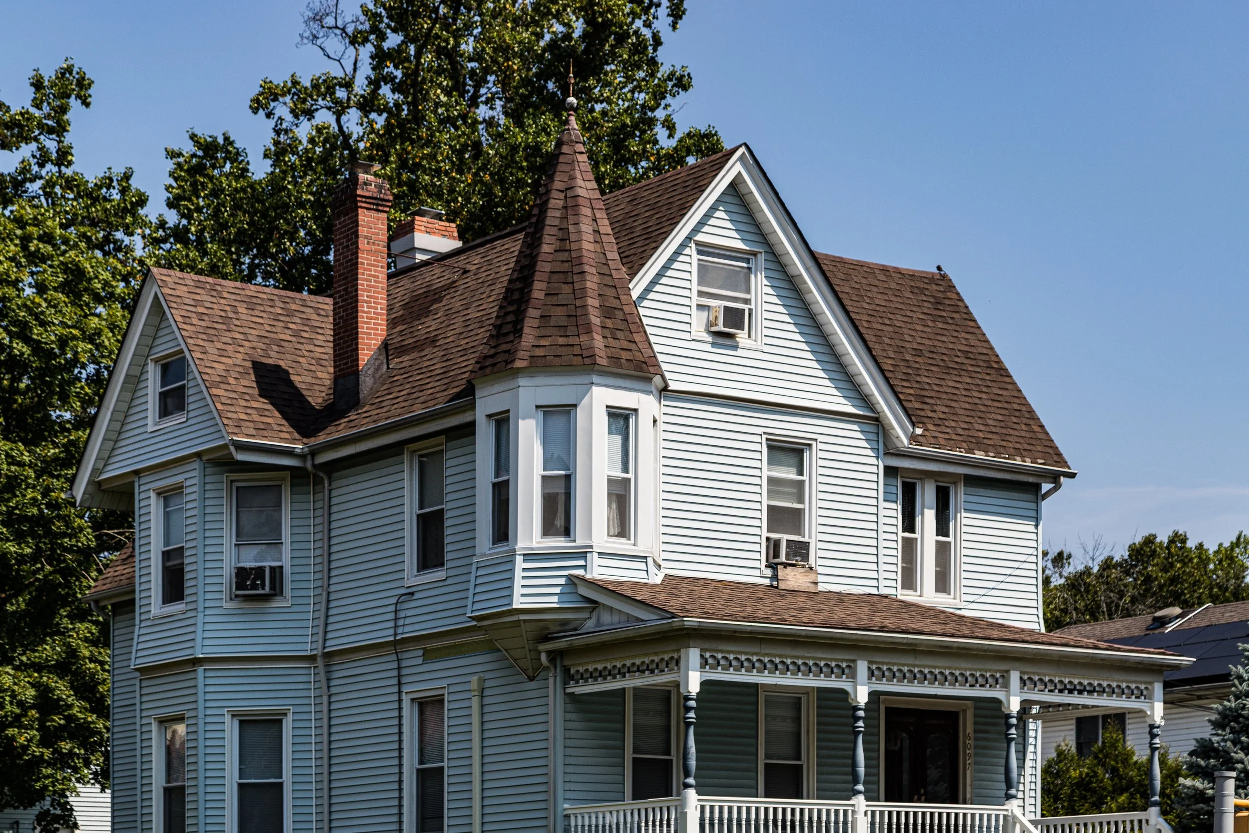 A large Victorian-style house with white siding, multiple gabled roofs, and a turret with a conical roof. The house has several double-hung windows, some with air conditioning units installed. There is a porch with decorative trim and columns, and a chimney is visible on one side. Bright daylight and a clear blue sky are present.