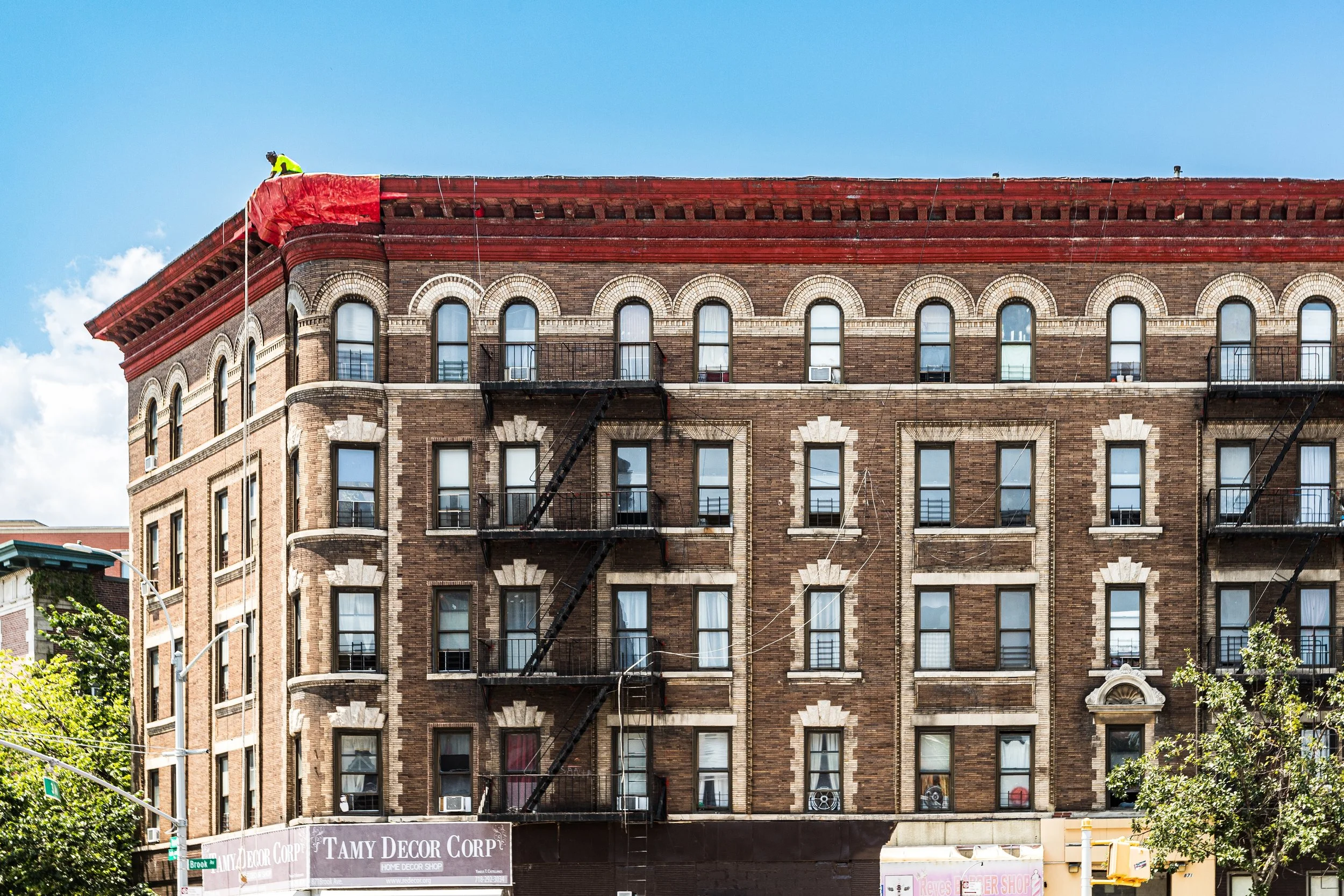 A multi-story brick building with fire escapes on the front. A construction worker is working at the roof, which is covered in red tarps. The building has several windows and decorative brickwork. Trees and street signs are visible in the foreground.