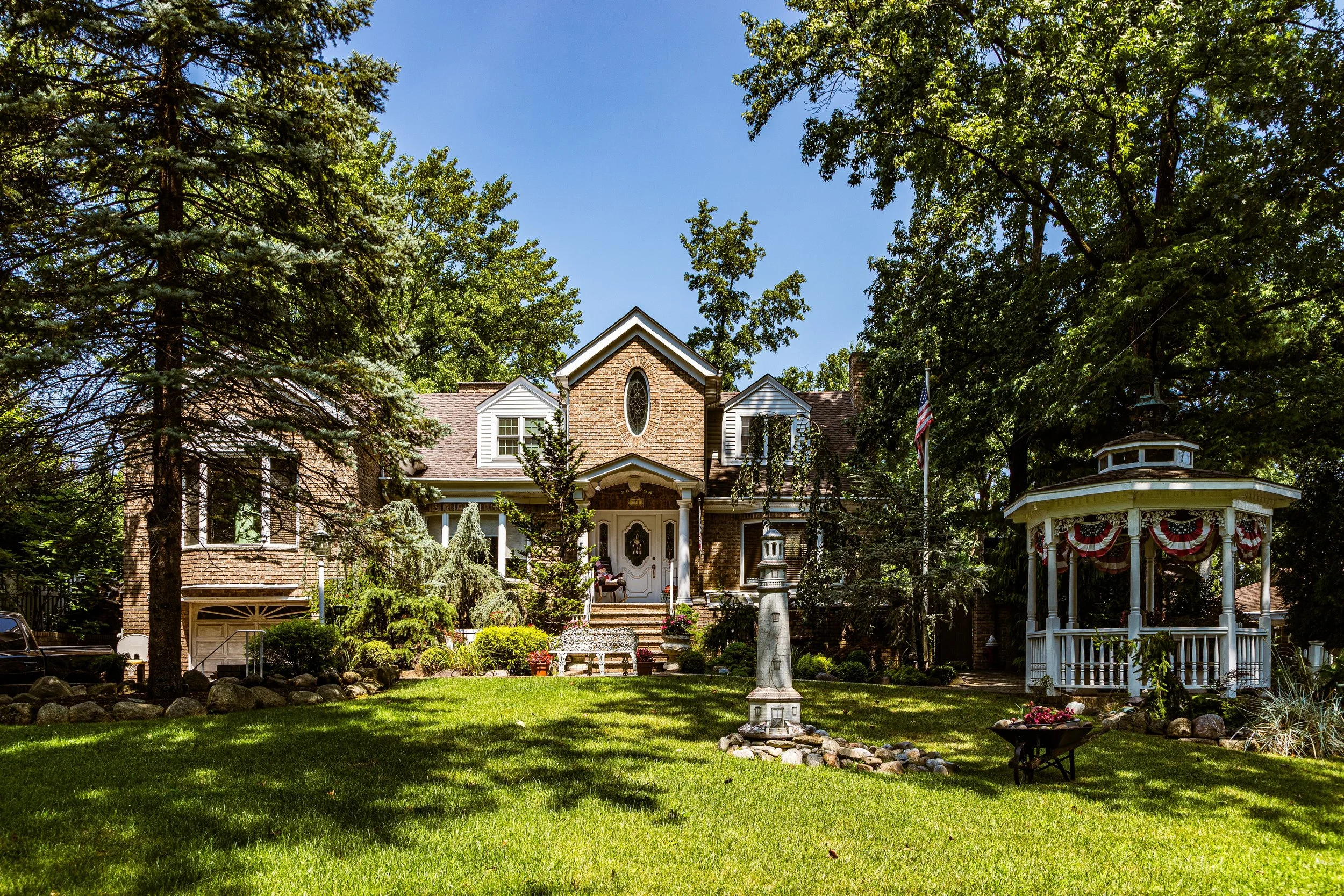 Large brick house with a porch, surrounded by tall trees and a well-maintained lawn. Features a white gazebo decorated with red, white, and blue bunting, and a flagpole with an American flag. A white fountain is in the center of the lawn.