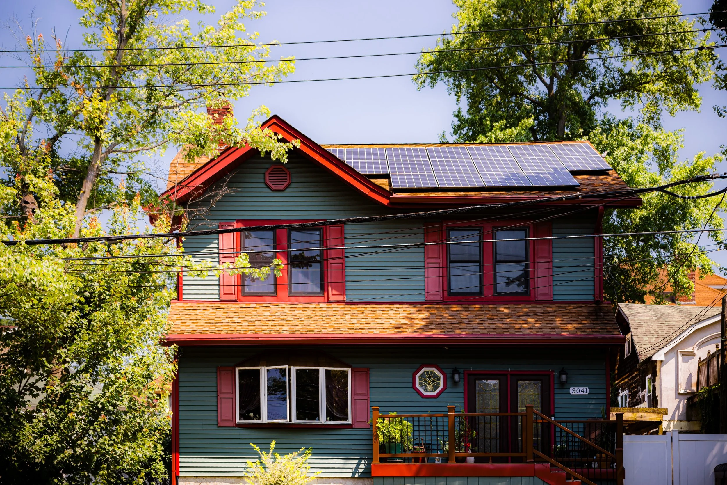 Colorful two-story house with green and red exterior, solar panels on roof, surrounded by trees, with a front porch and a white fence.