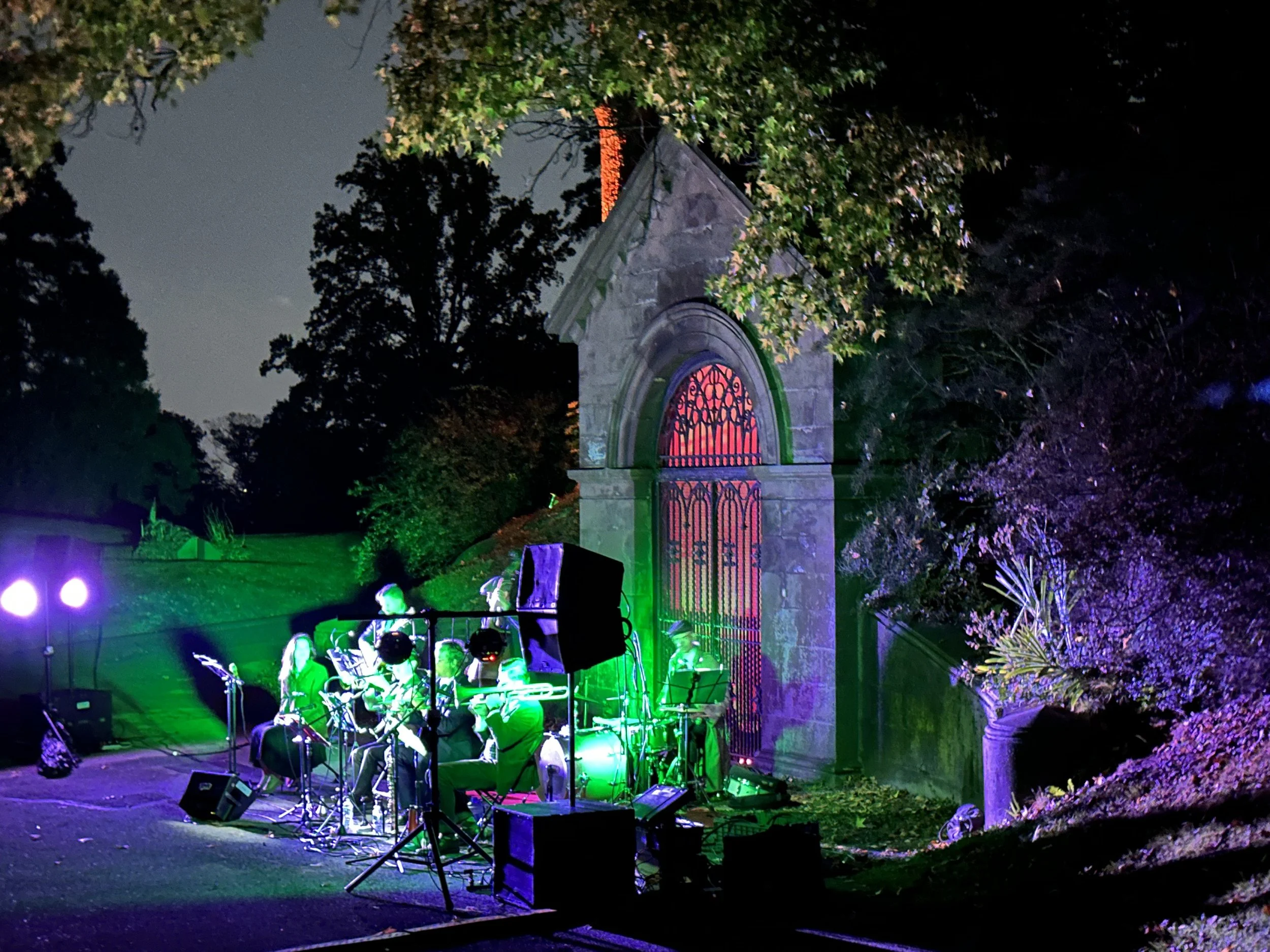 A nighttime outdoor concert with musicians playing in front of an old stone building with a large arched window and iron gate, illuminated by colorful green, purple, and pink lights.