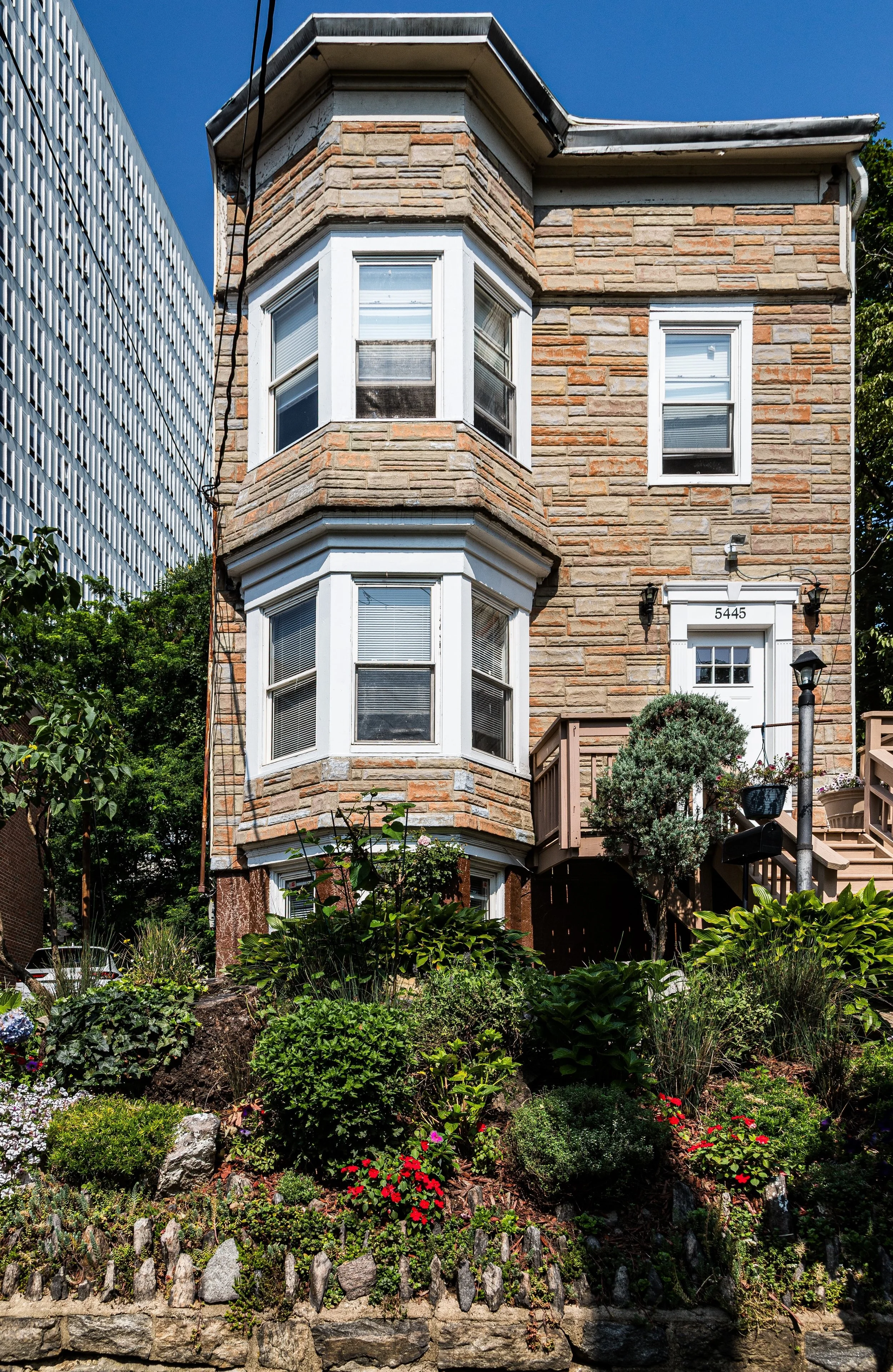 A three-story brick house with a bay window on each floor, a white door with the number 5445, and a front garden with various plants and flowers, including red, white, and green foliage.