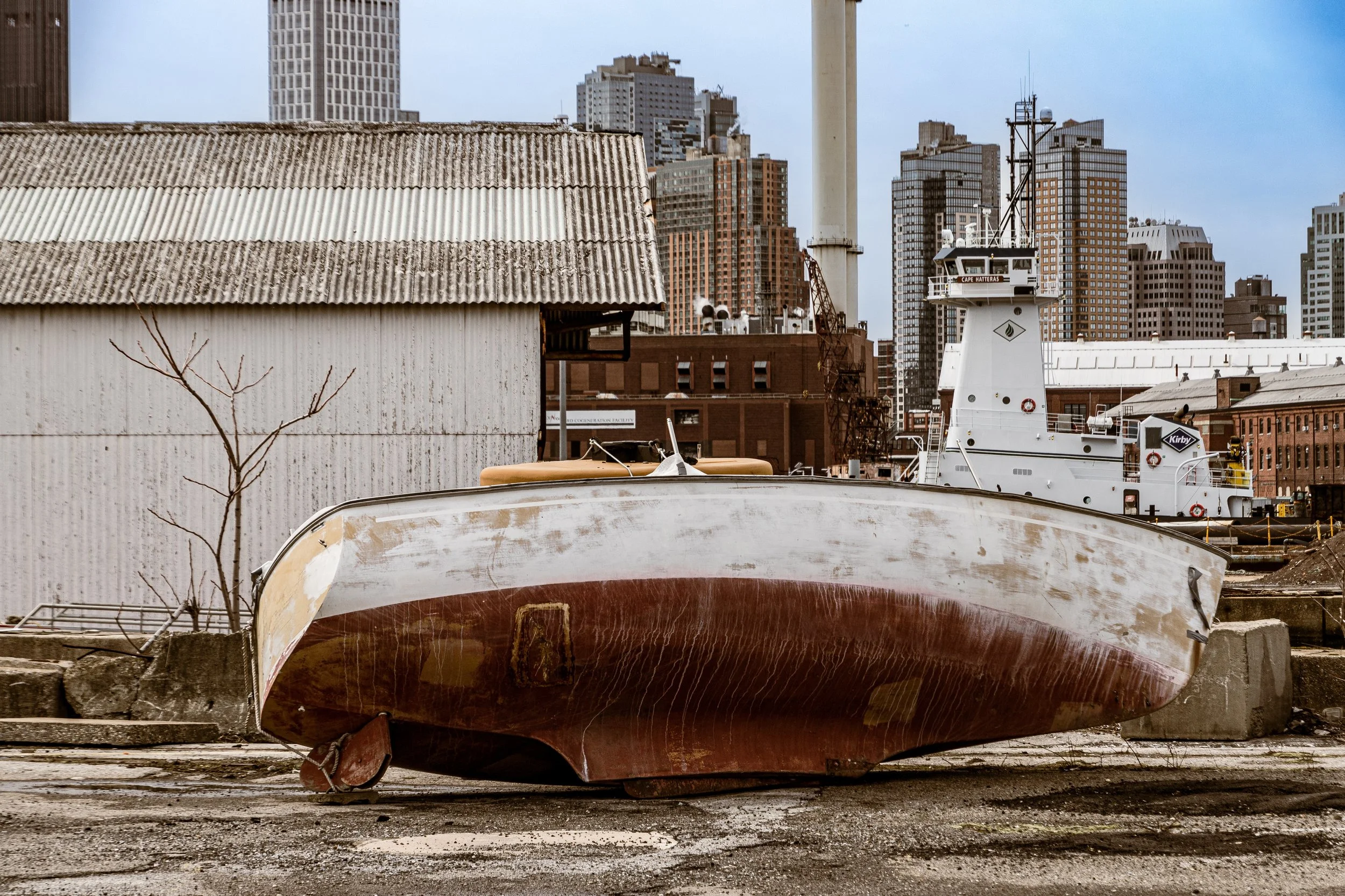 An old, weathered boat resting on a dock with an urban cityscape in the background, featuring tall buildings and a crane.