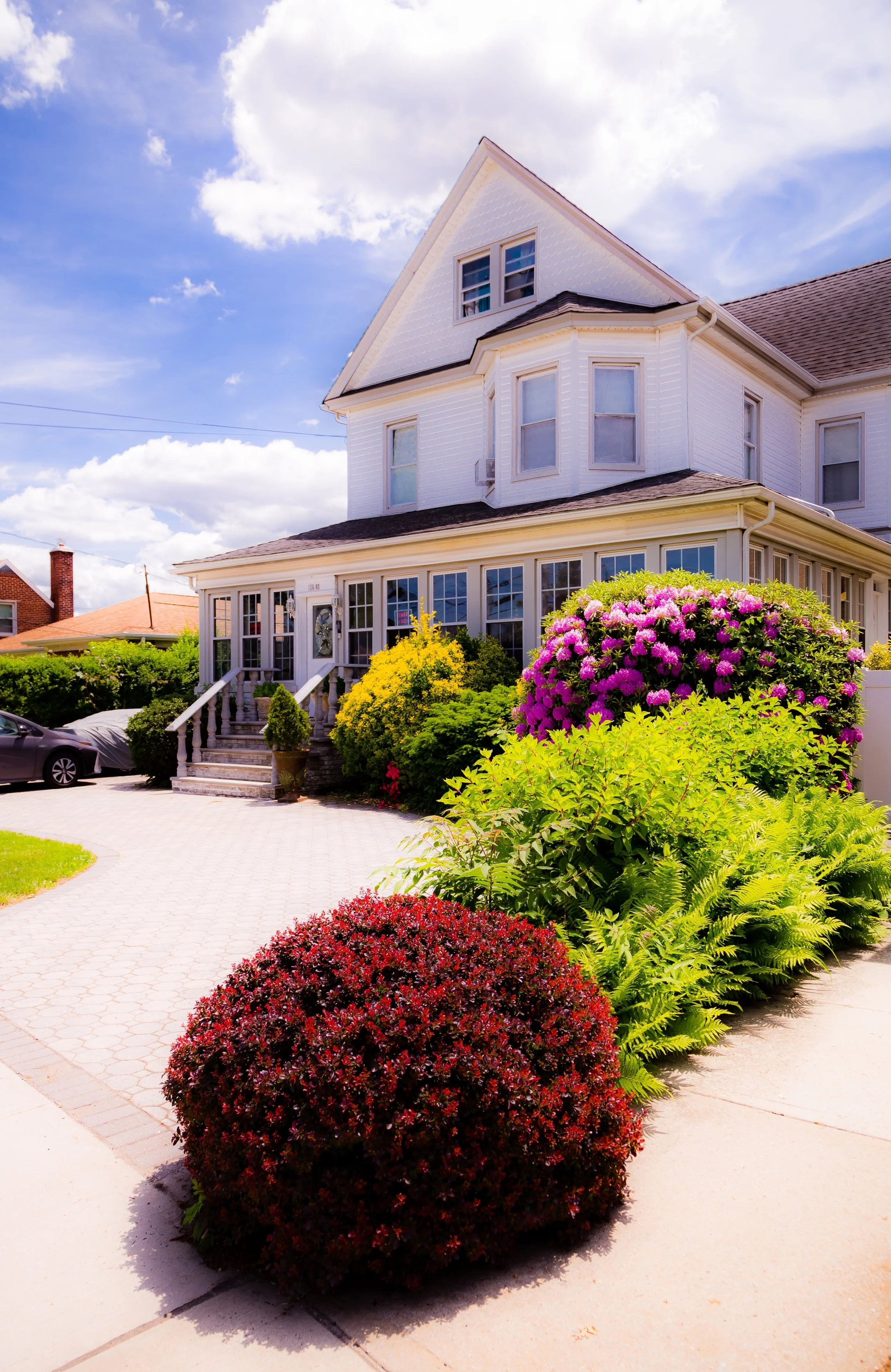 A large white Victorian-style house with a front porch and a garden filled with colorful flowering shrubs and bushes, under a partly cloudy sky.