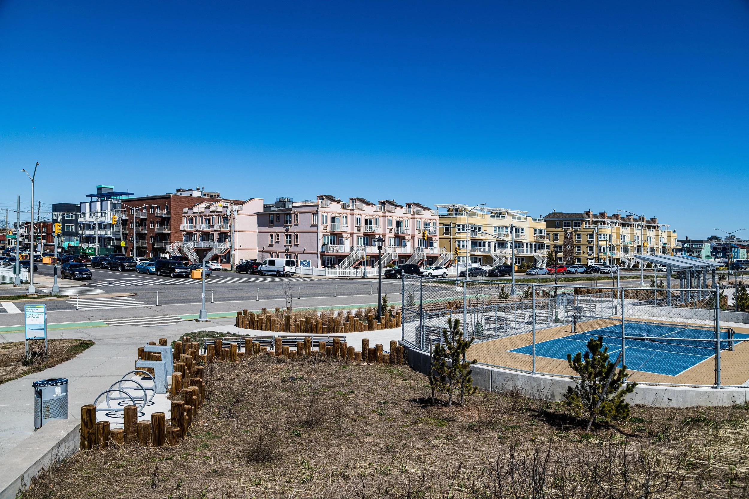 A clear day view of a residential area with pink and yellow multi-story buildings, a parking lot filled with cars, a tennis court with a chain-link fence, and a landscaped area with small trees and flower beds.