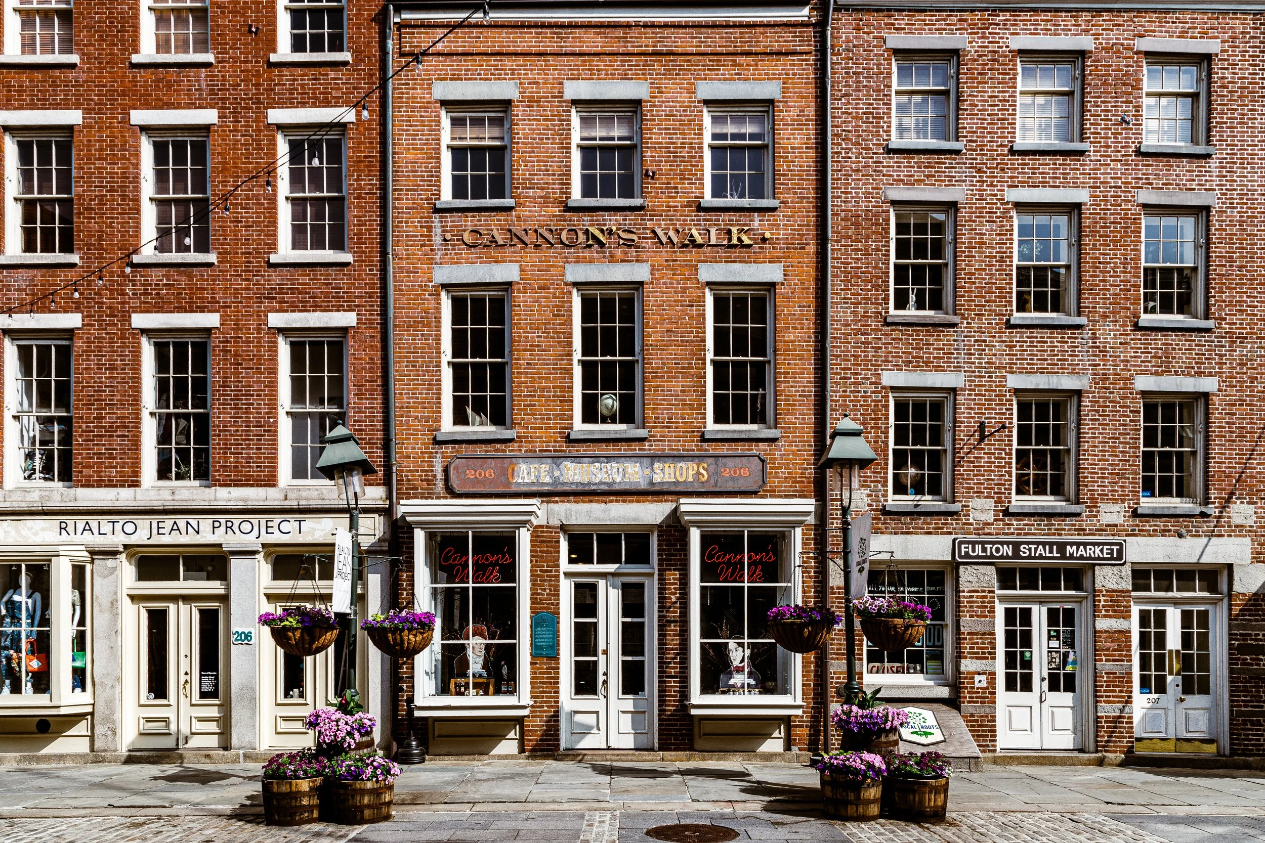 A row of historic brick buildings on a city street with store windows and hanging flower baskets.