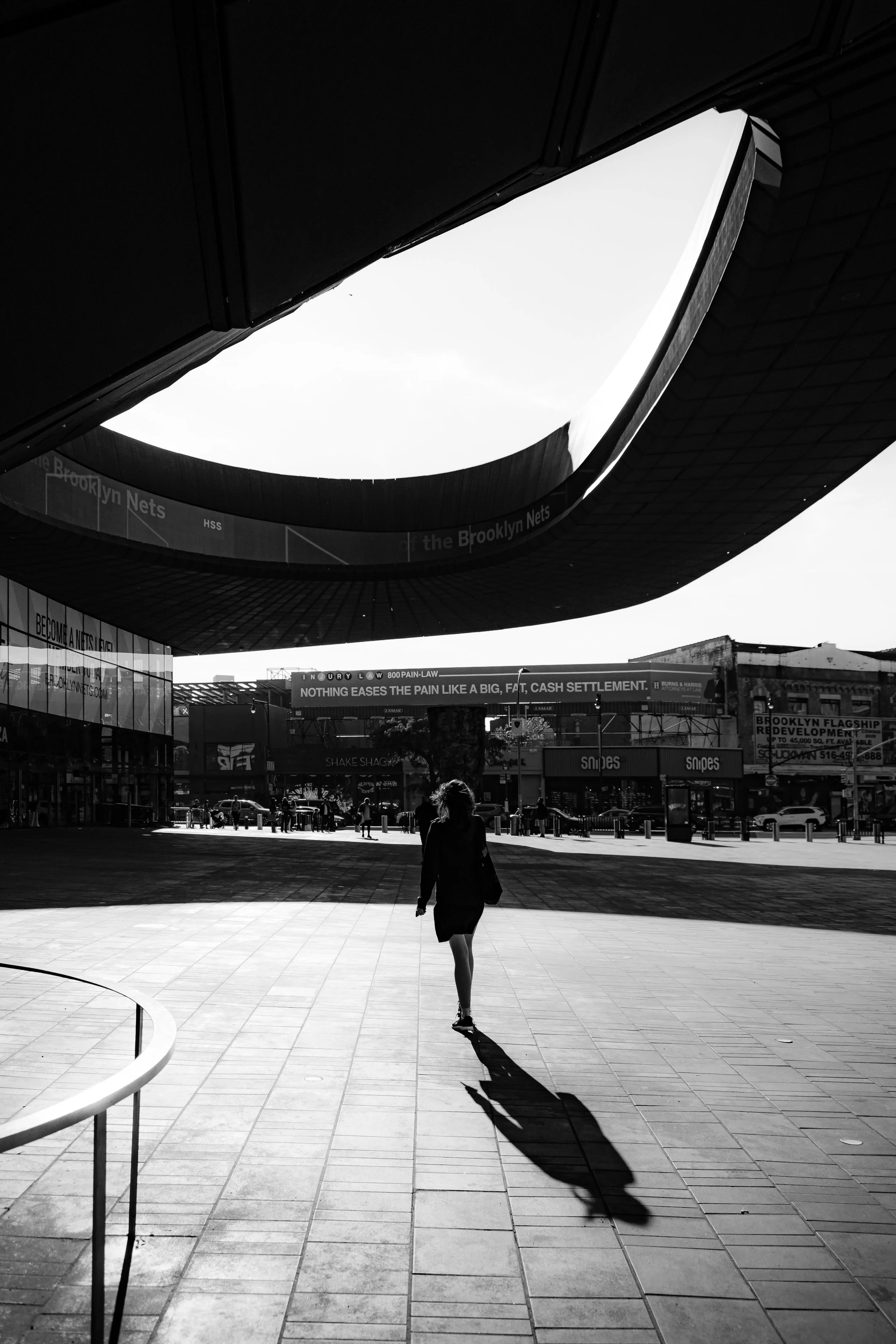 A woman walking in an urban plaza with modern architecture overhead and signs for Brooklyn Nets in the background, in black and white.