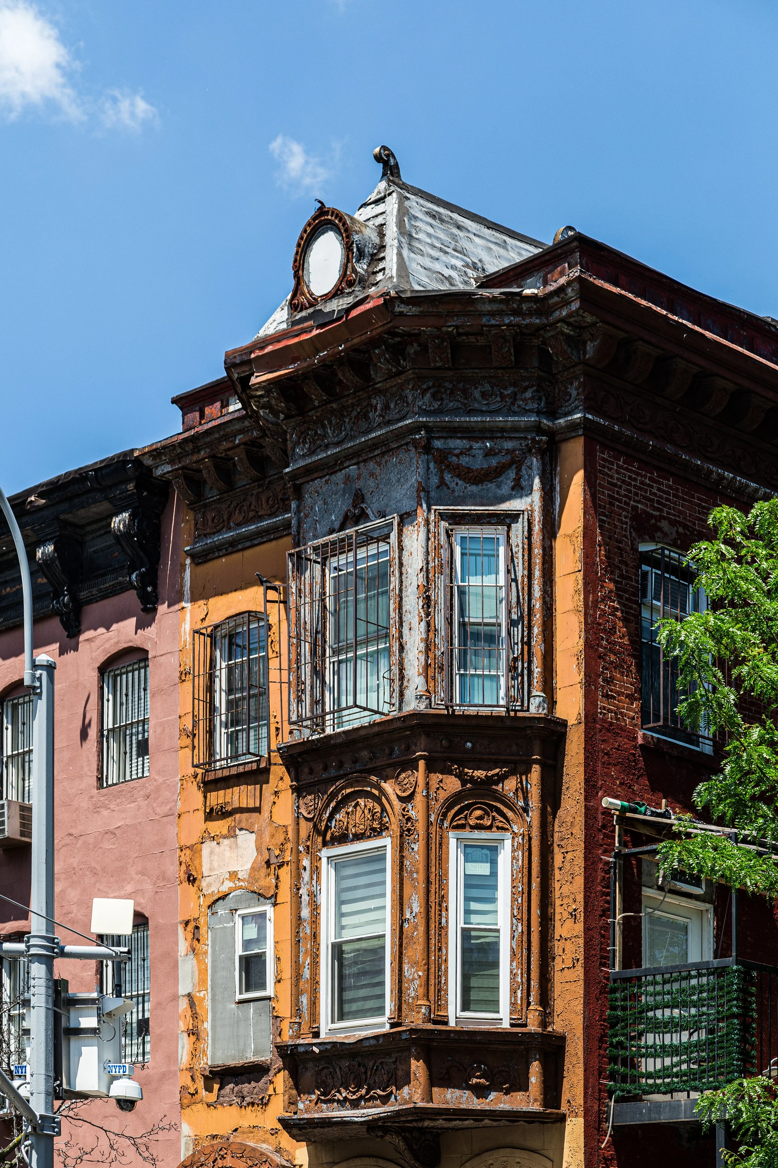 Old, weathered building with ornate bay windows, rusted iron bars on windows, and peeling paint, under a blue sky.